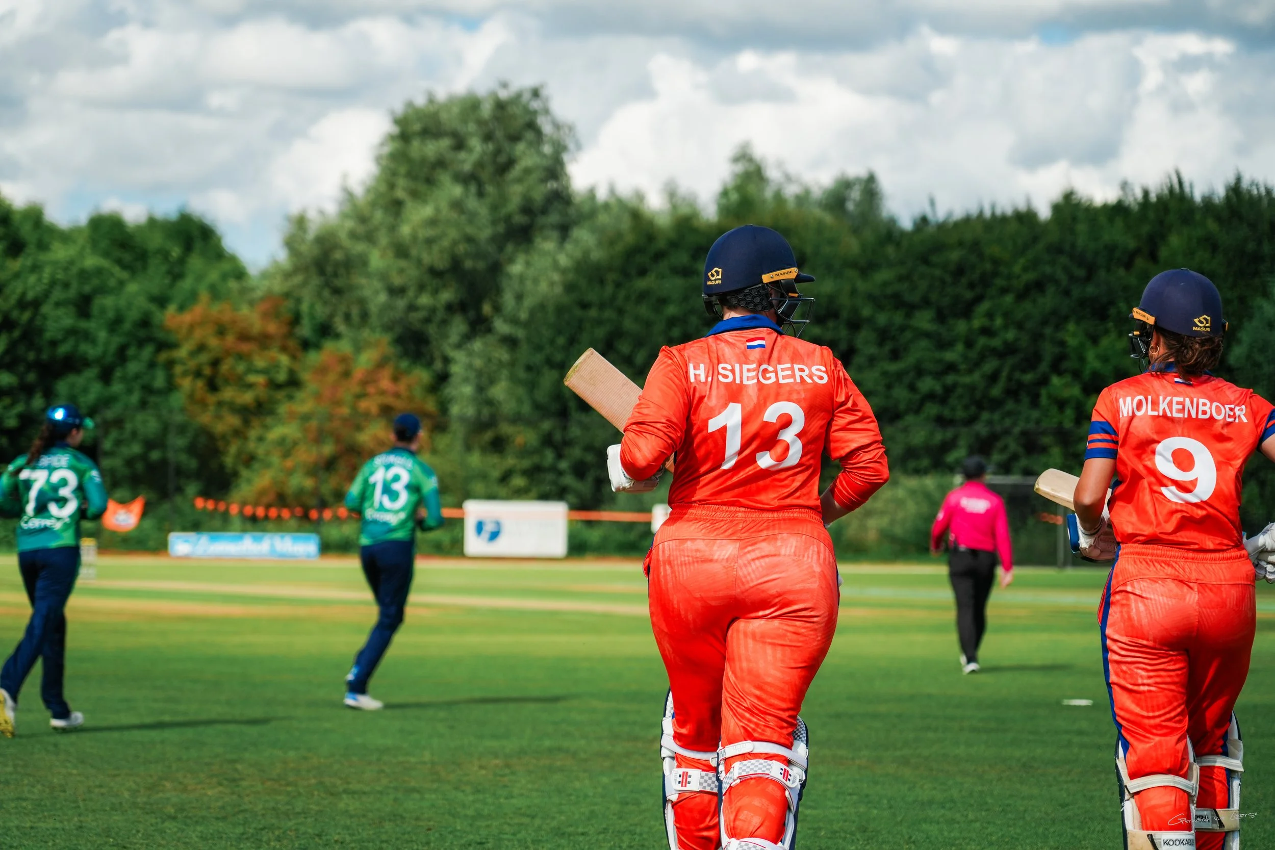 Female cricket players in orange uniforms with helmets, holding bats, walking on the field during a game, with other players in green uniforms and an umpire in the background, lush greenery, cloudy sky.