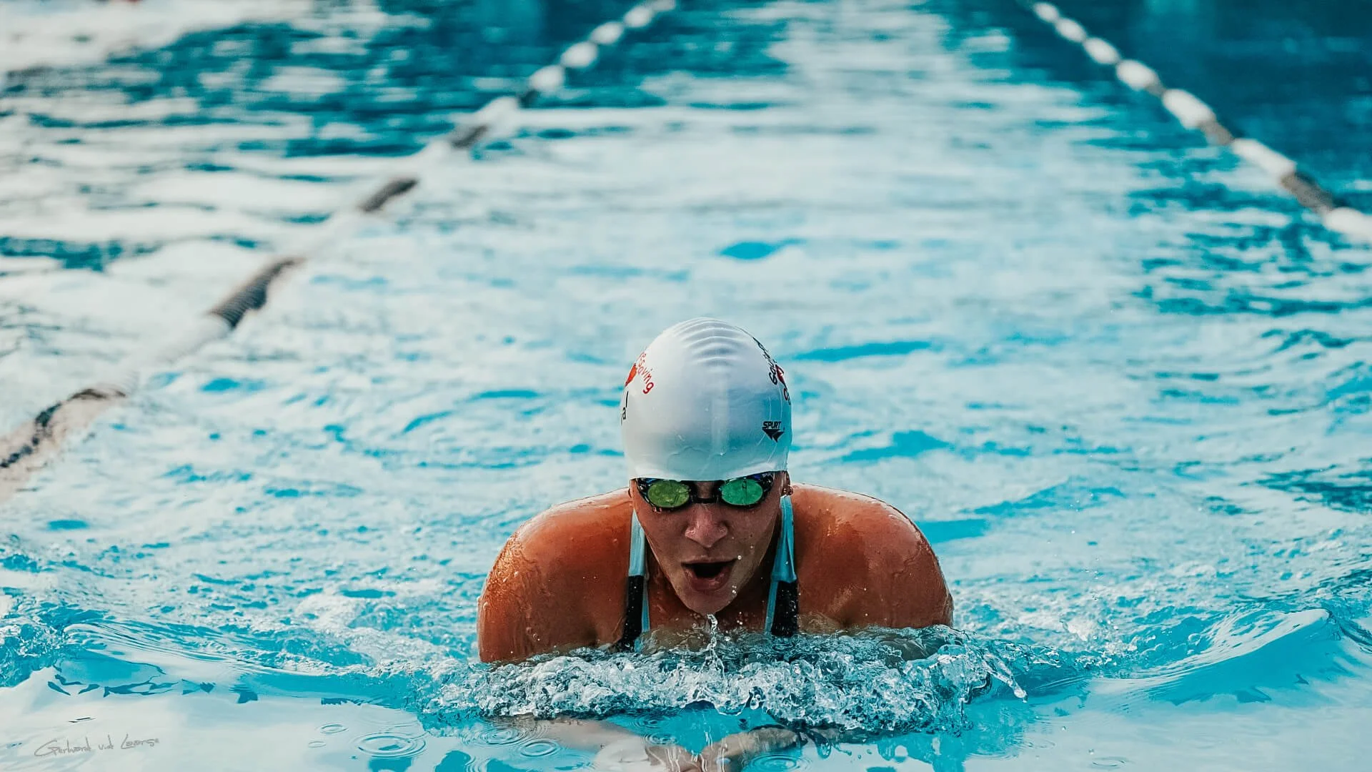Swimmer in a pool wearing a white swim cap and goggles, with arms extended forward and face in the water, swimming butterfly stroke.
