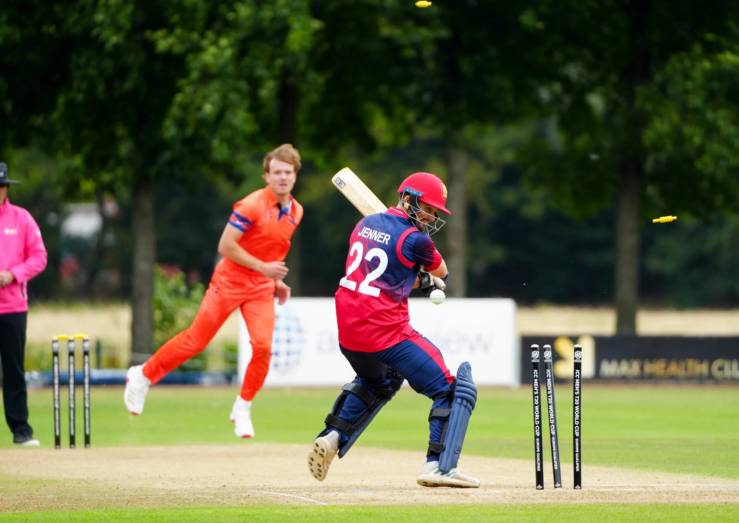 A women's cricket player in red and blue uniform, number 22, batting in a cricket match. A male player in an orange uniform is running nearby, and there are three wickets on the pitch. The game is outdoors on a grassy field with trees in the backgrou