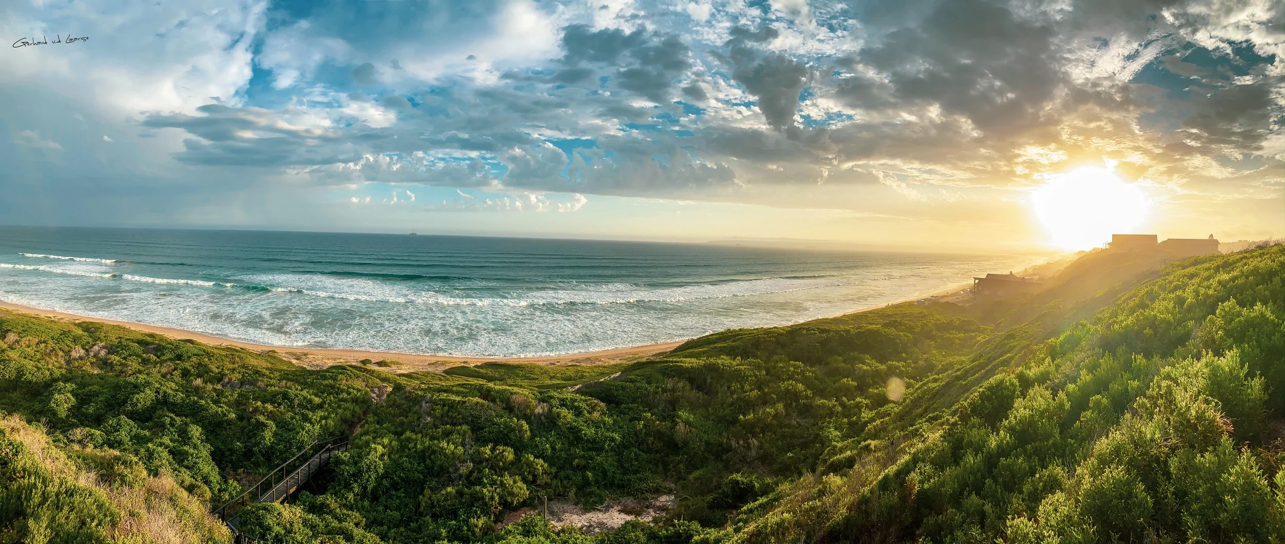 Sunset over the ocean viewed from a sandy beach with green bushes and houses on a hill.