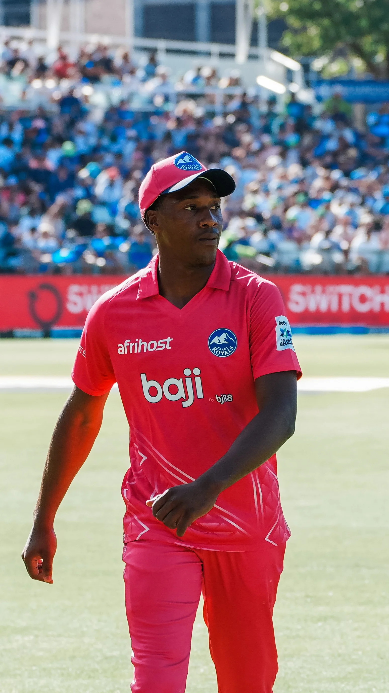 Cricket player in a red jersey and cap on the field during a match with spectators in the background.