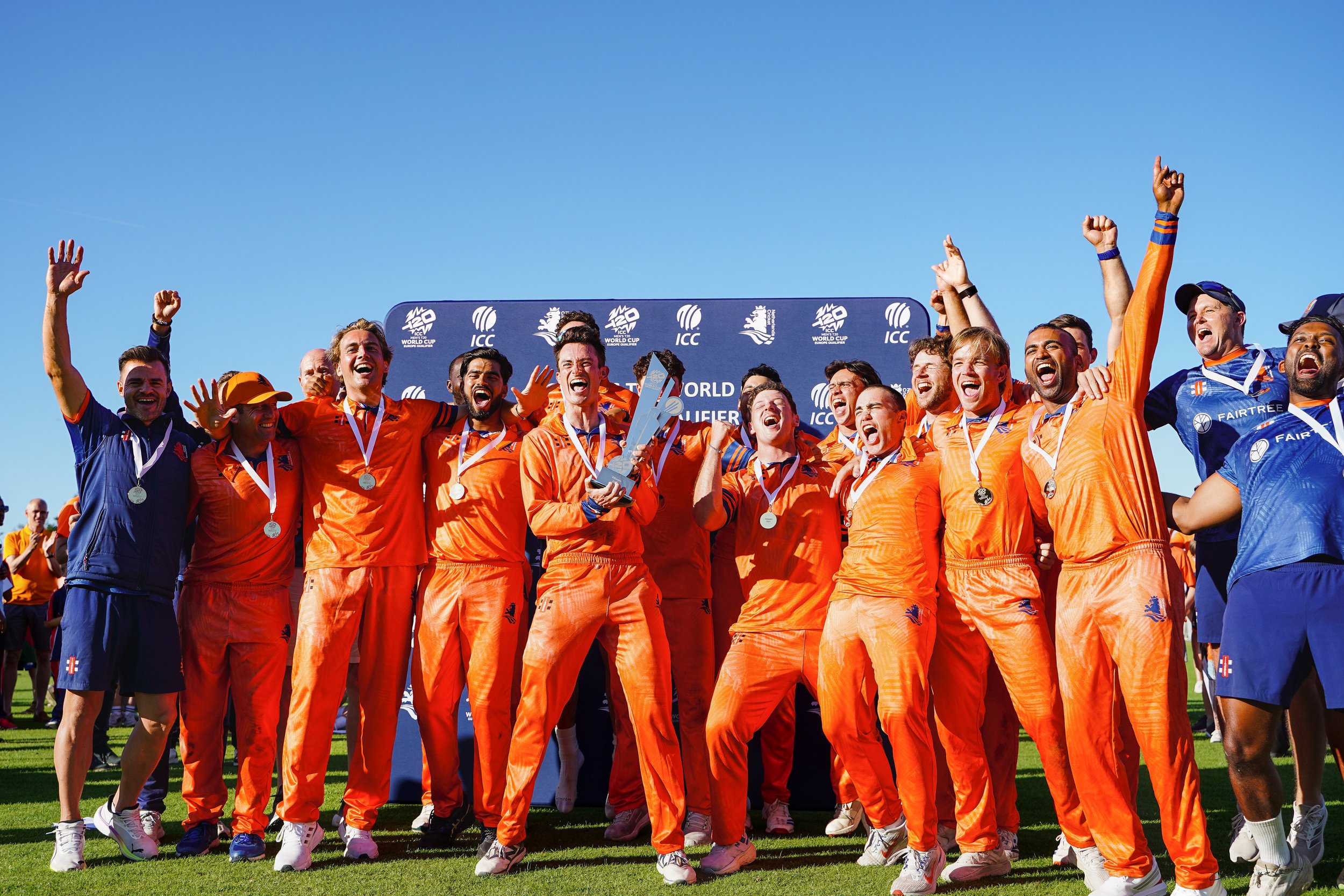 A cricket team celebrating with medals and trophies on a field, wearing orange and blue uniforms, with a backdrop displaying logos and text indicating a world cricket tournament.