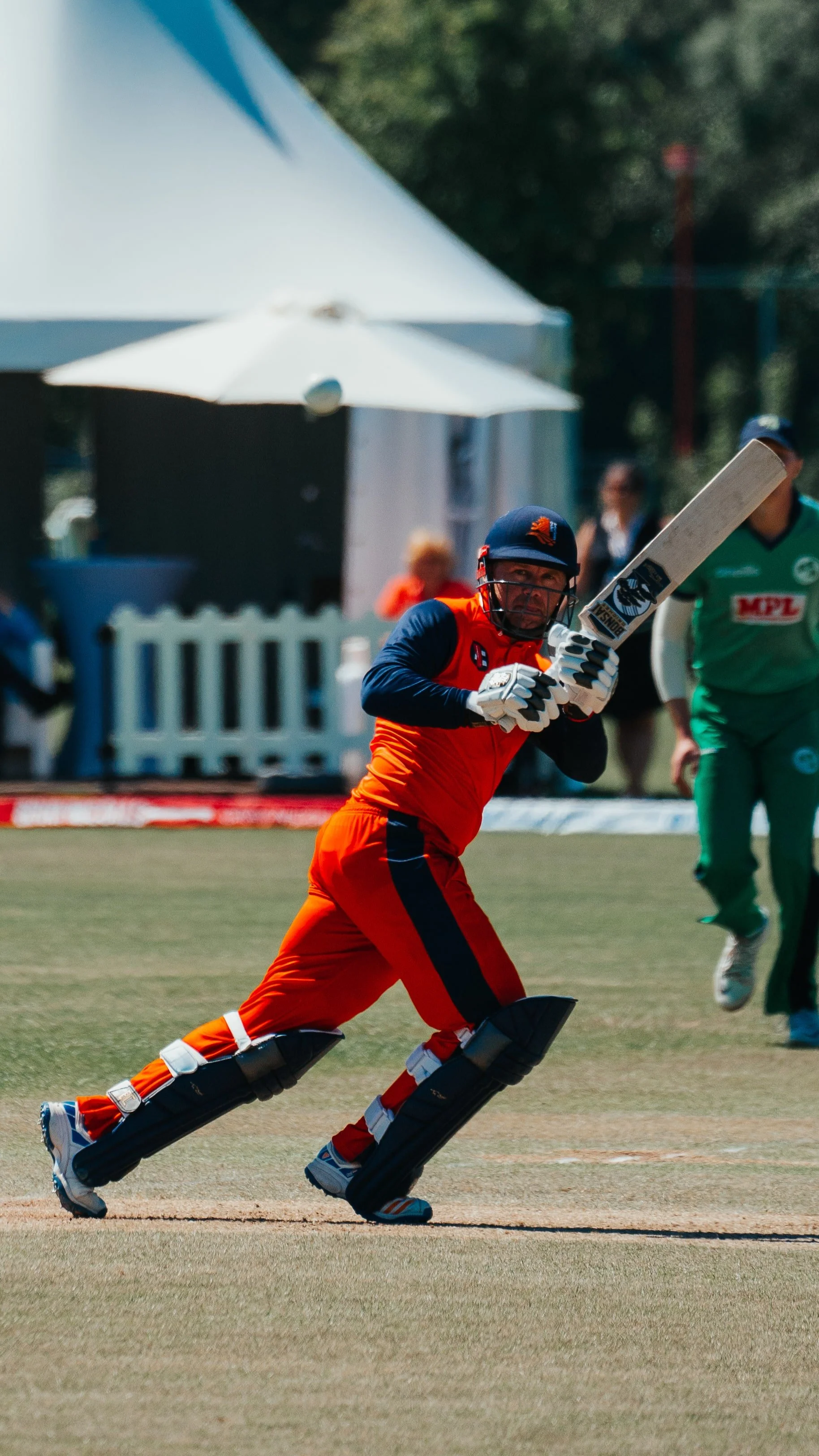 A cricket player in orange and navy uniform, wearing a helmet and padded leg guards, is batting on a cricket field during daytime. He appears to be running or preparing to hit the ball.
