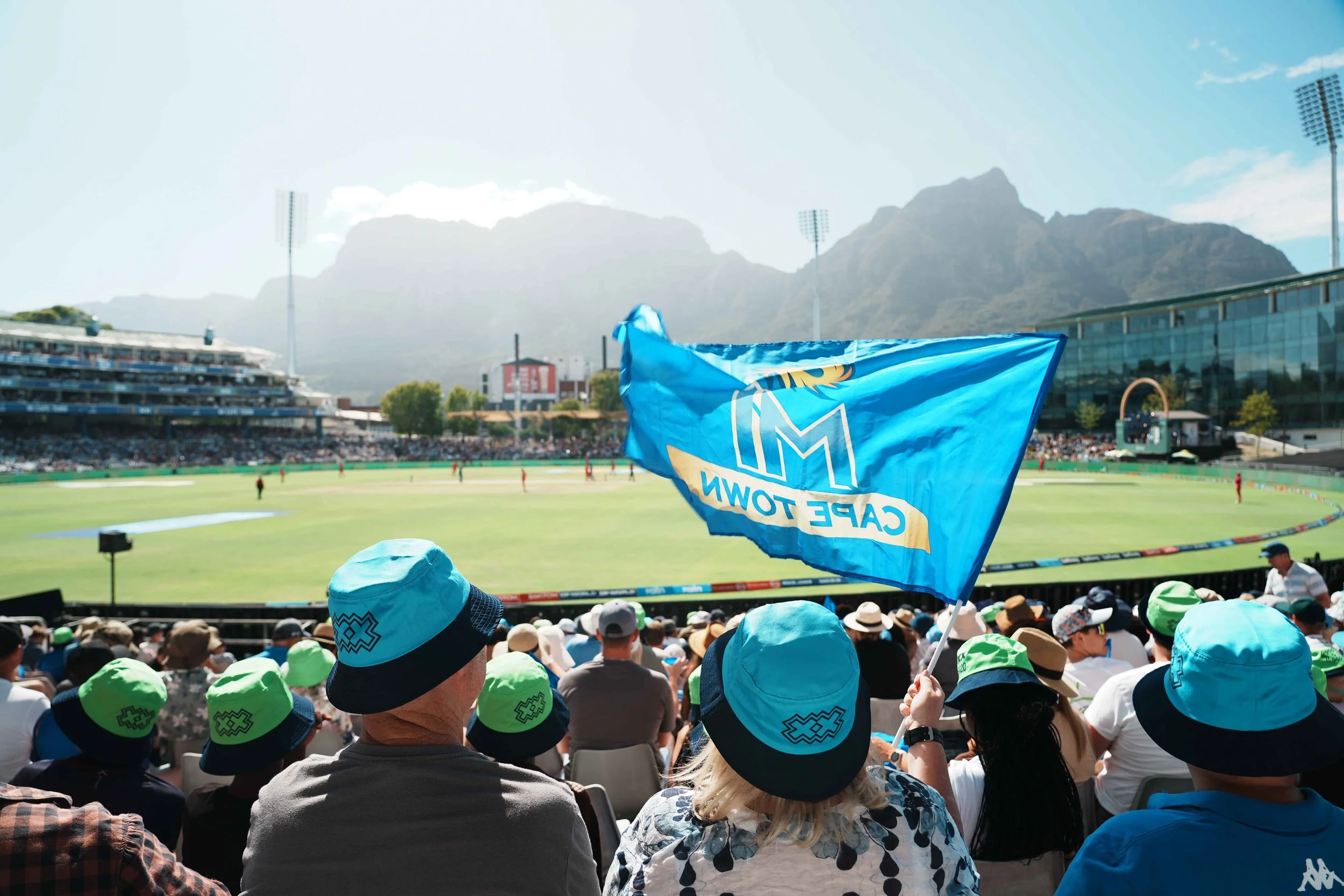 Crowd watching a cricket game at a stadium with a blue Cape Town flag flying in the foreground and mountains in the background.