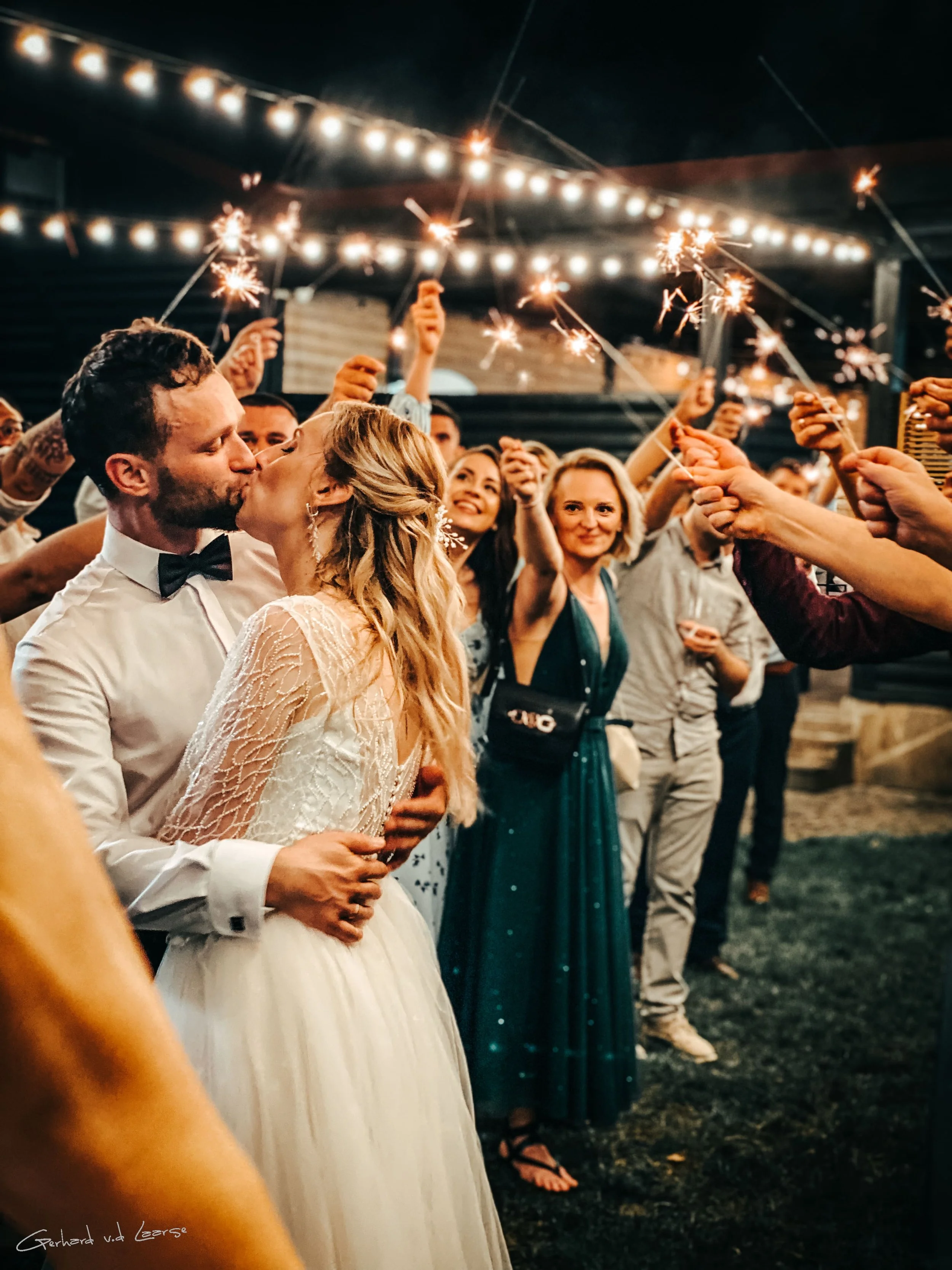 A bride and groom kiss during their wedding reception as friends hold sparklers in the background, under string lights at night.