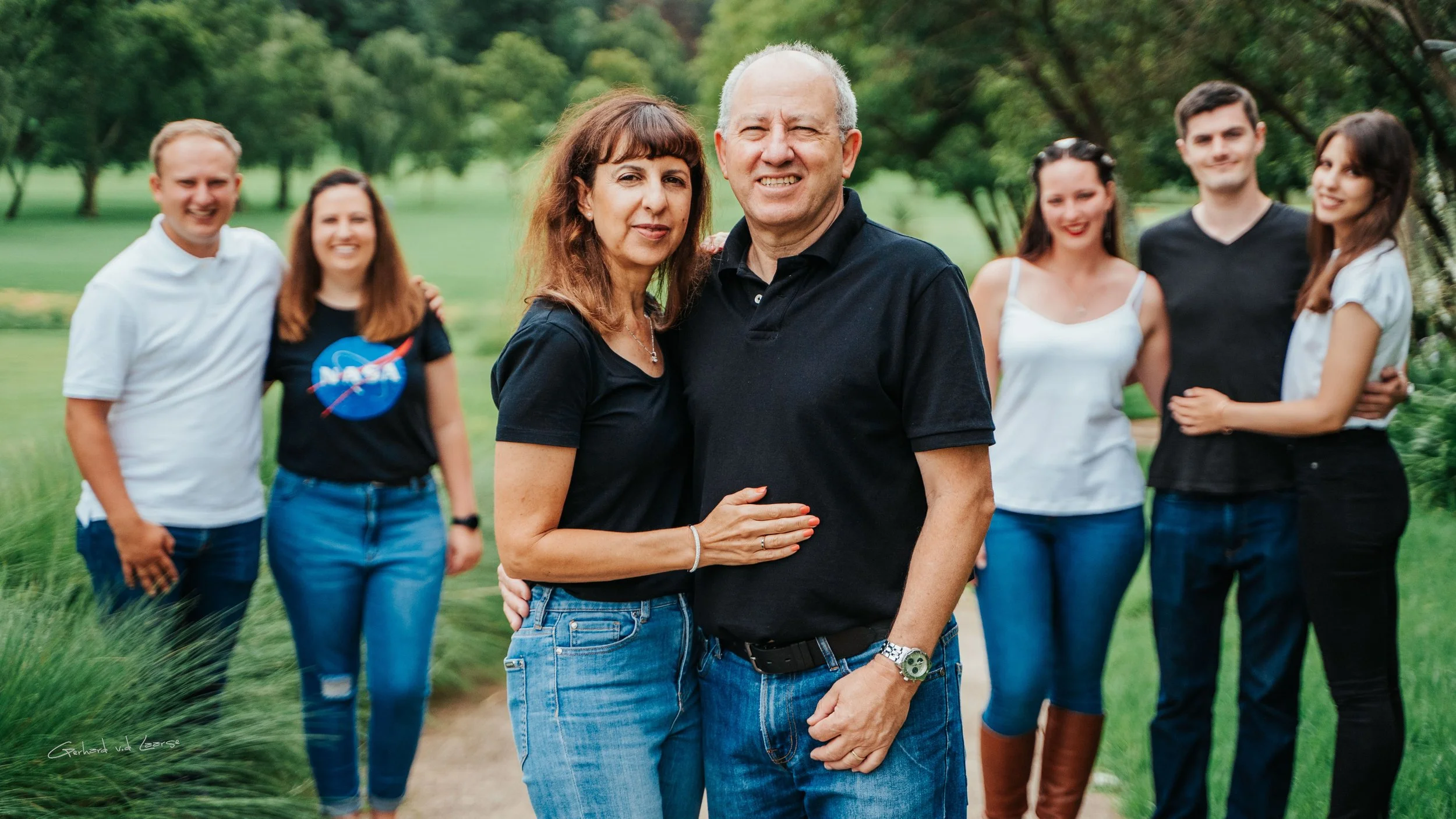A family portrait of seven people standing outdoors in a park with green grass and trees in the background, smiling, with two people in the front and five behind.
