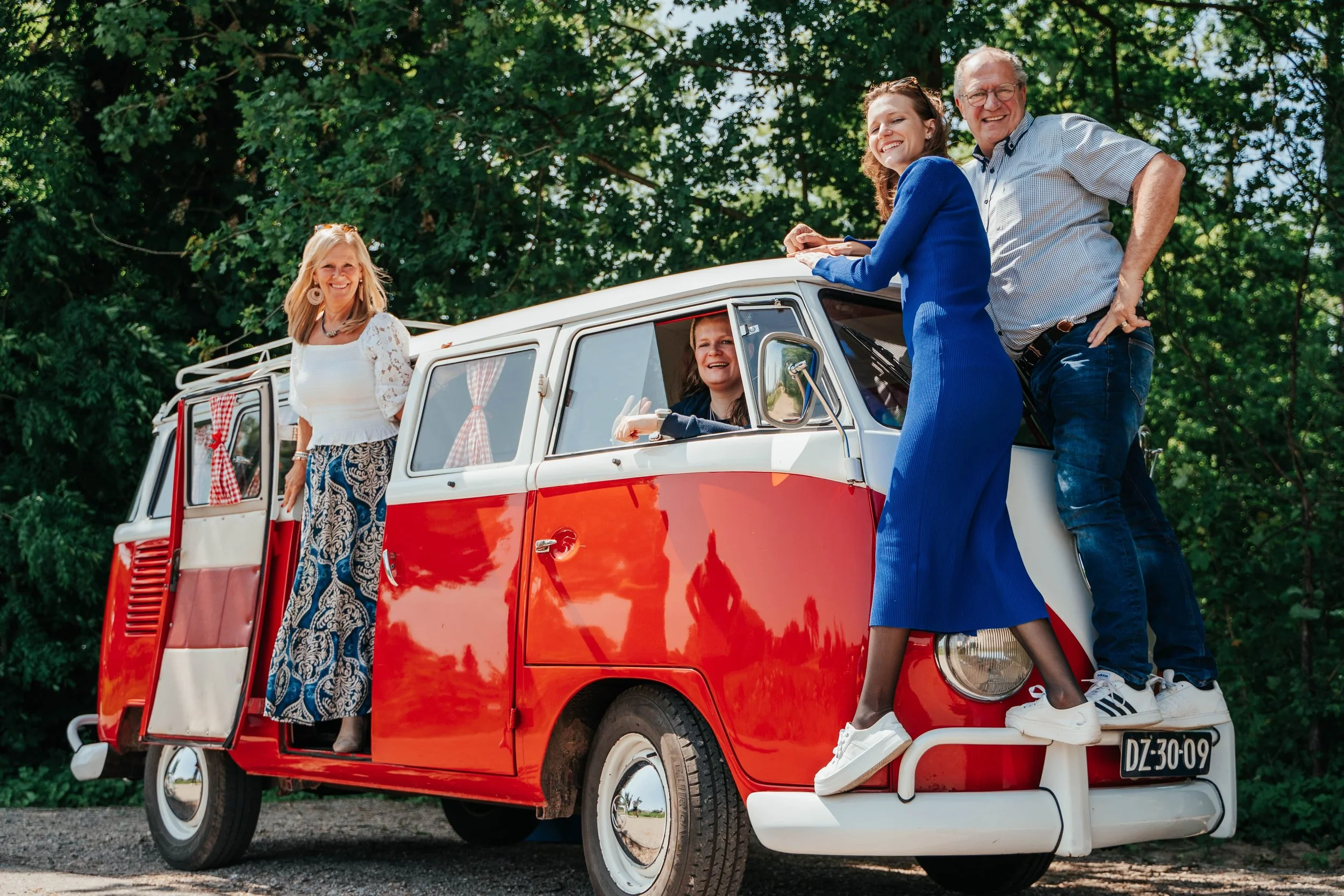 Four smiling women and one man enjoying a day outdoors with a red and white vintage van, two women are sitting on the front of the van, while two women are standing near the open door and the back of the van.