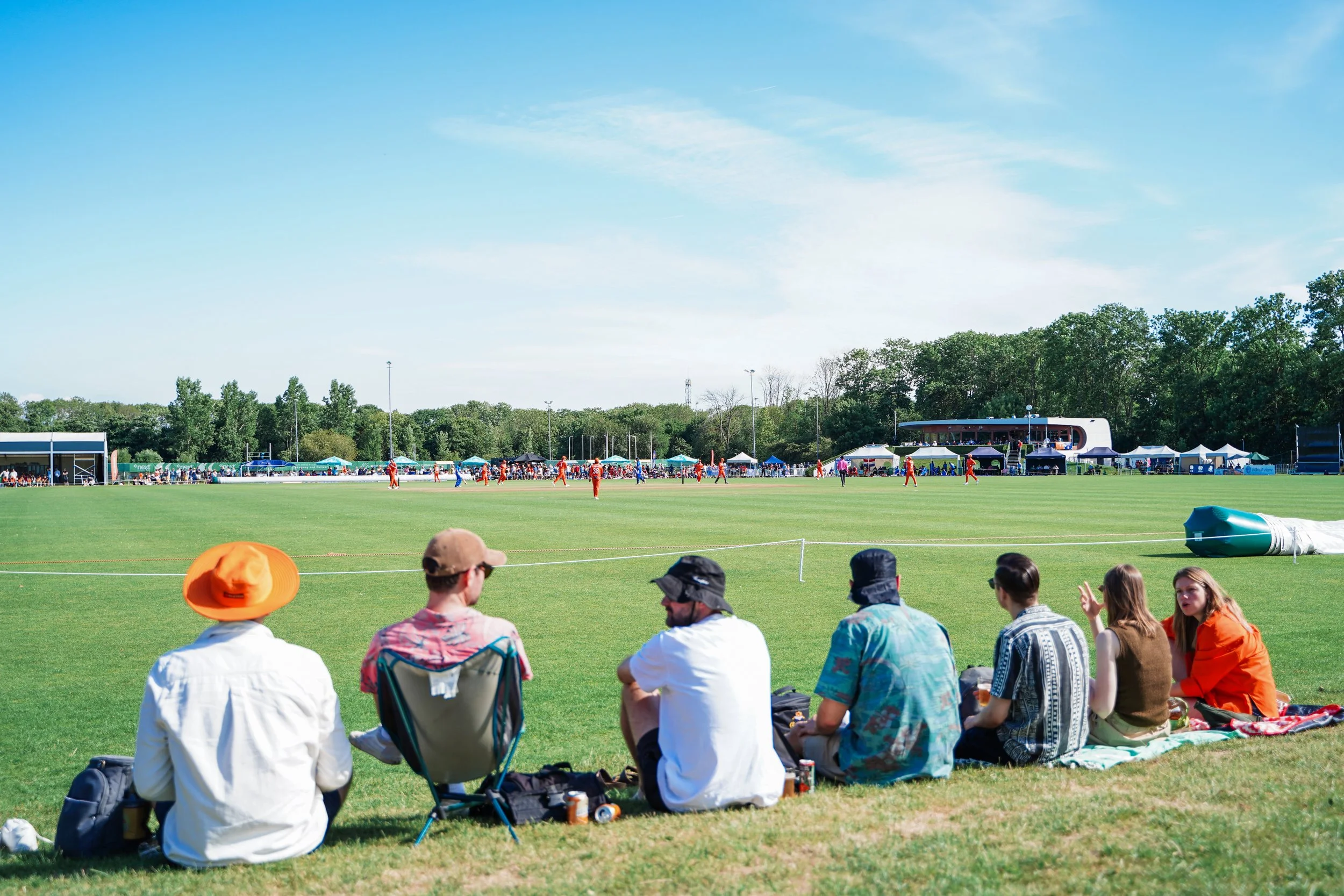People sitting on the grass watching a cricket match on a sunny day at a sports field with trees, tents, and spectators in the background.