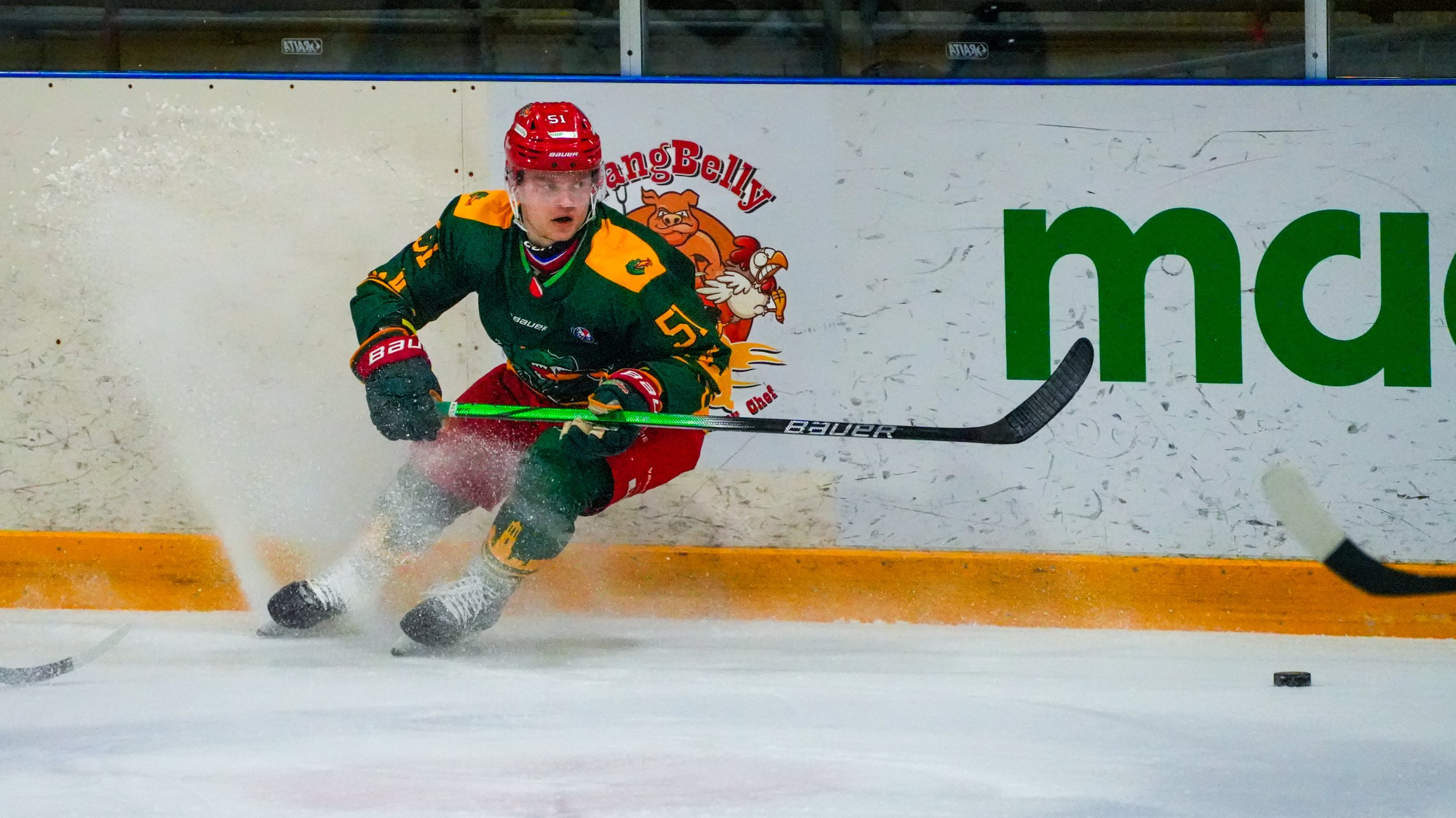 Ice hockey player in green and red uniform with a red helmet skating on ice with a hockey stick, near the boards with a logo, in an indoor rink.