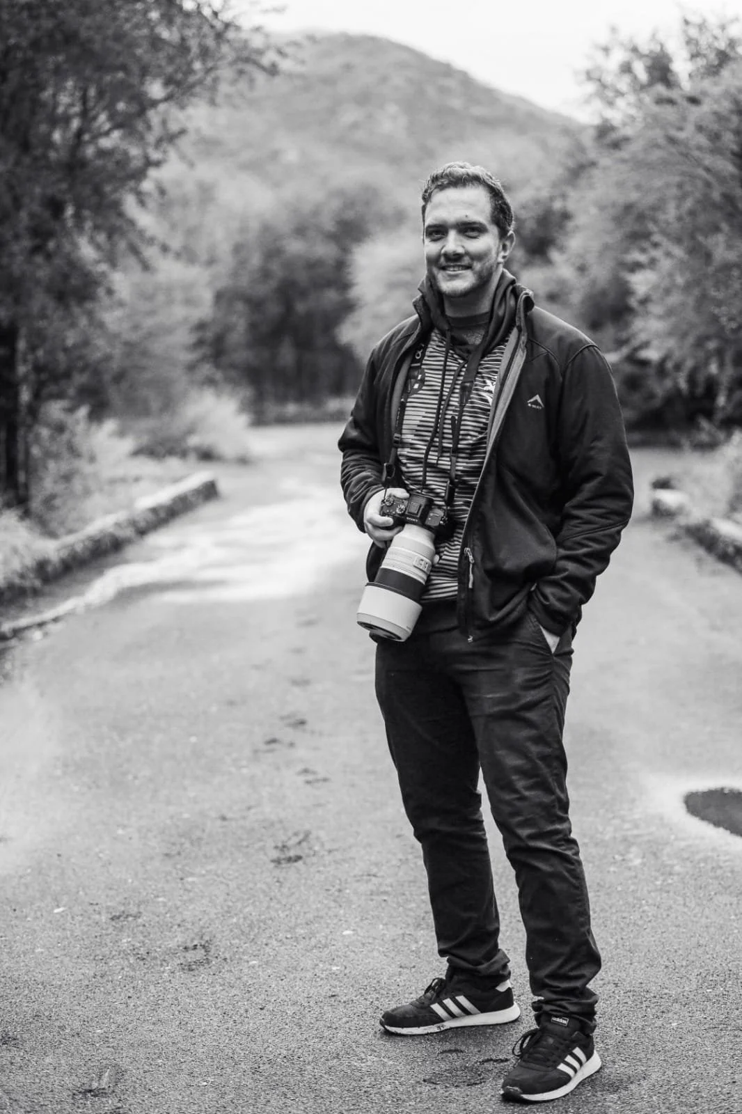 A man smiling outdoors on a path with a camera around his neck, surrounded by trees and natural scenery.