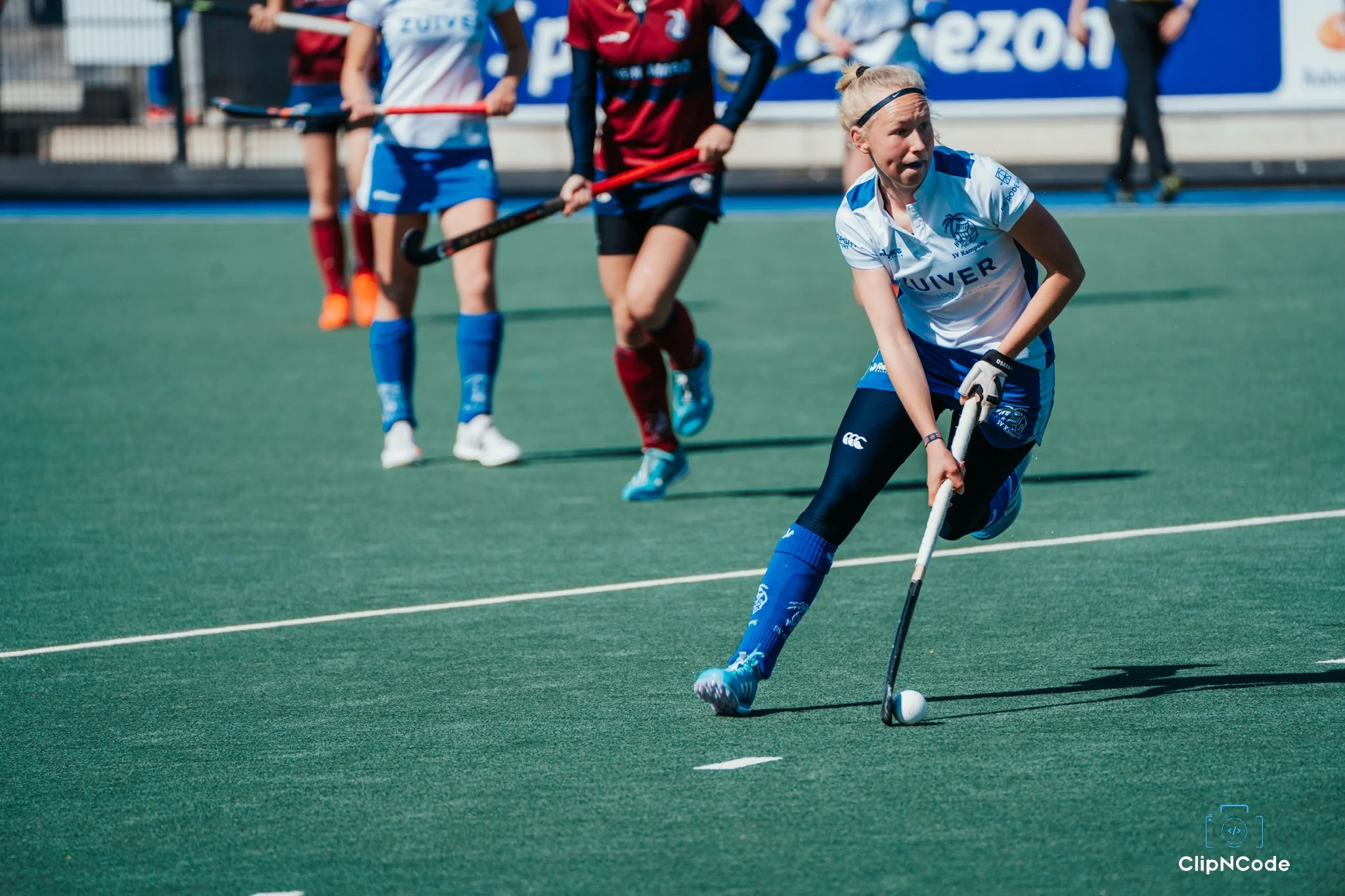 A field hockey player in a white and blue uniform controls the ball with her stick during a game, with other players in red and blue uniforms behind her on a green turf field.