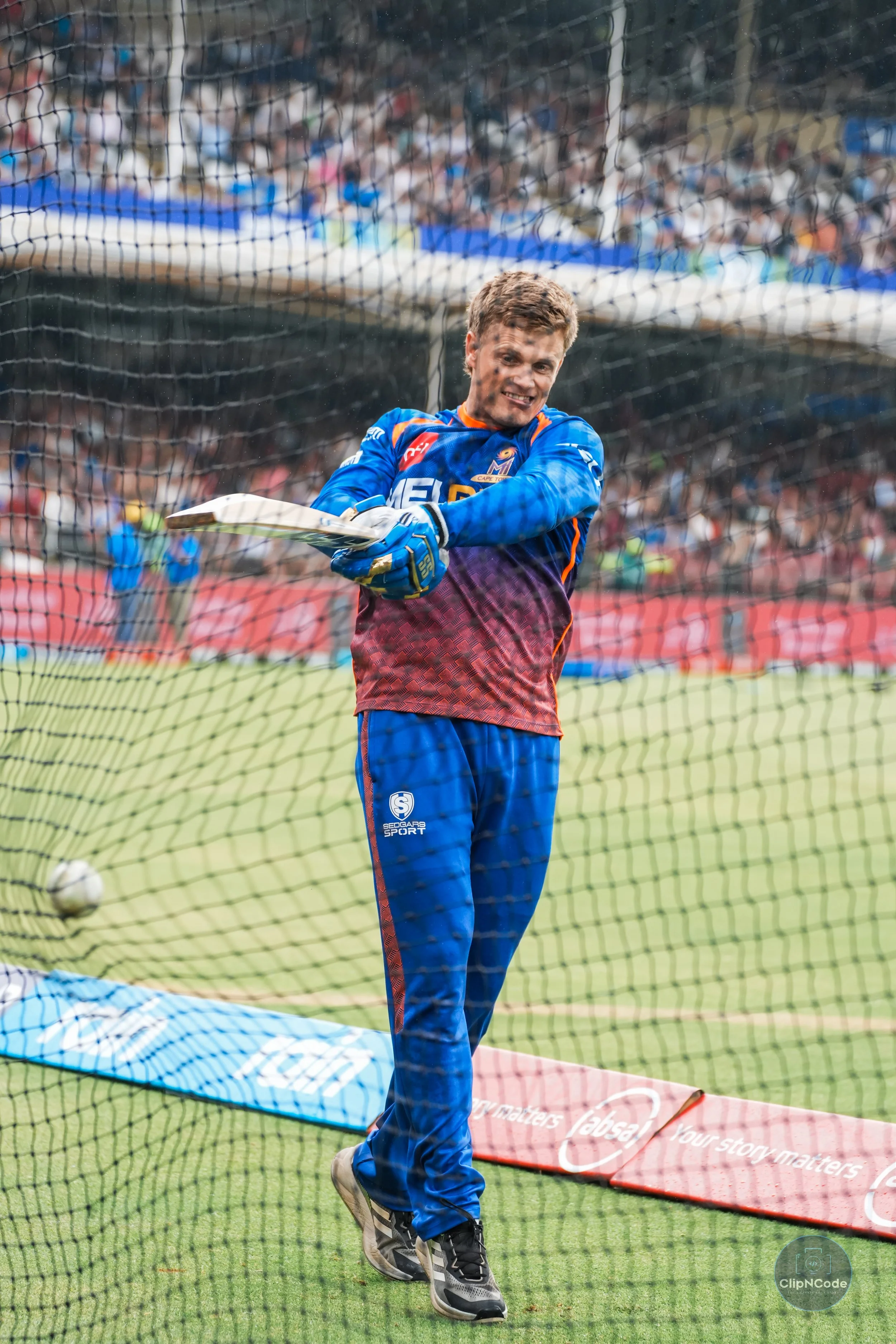 An athlete celebrating on a sports field, wearing blue and purple sportswear with gloves, and holding a hockey stick, standing behind a goal net.
