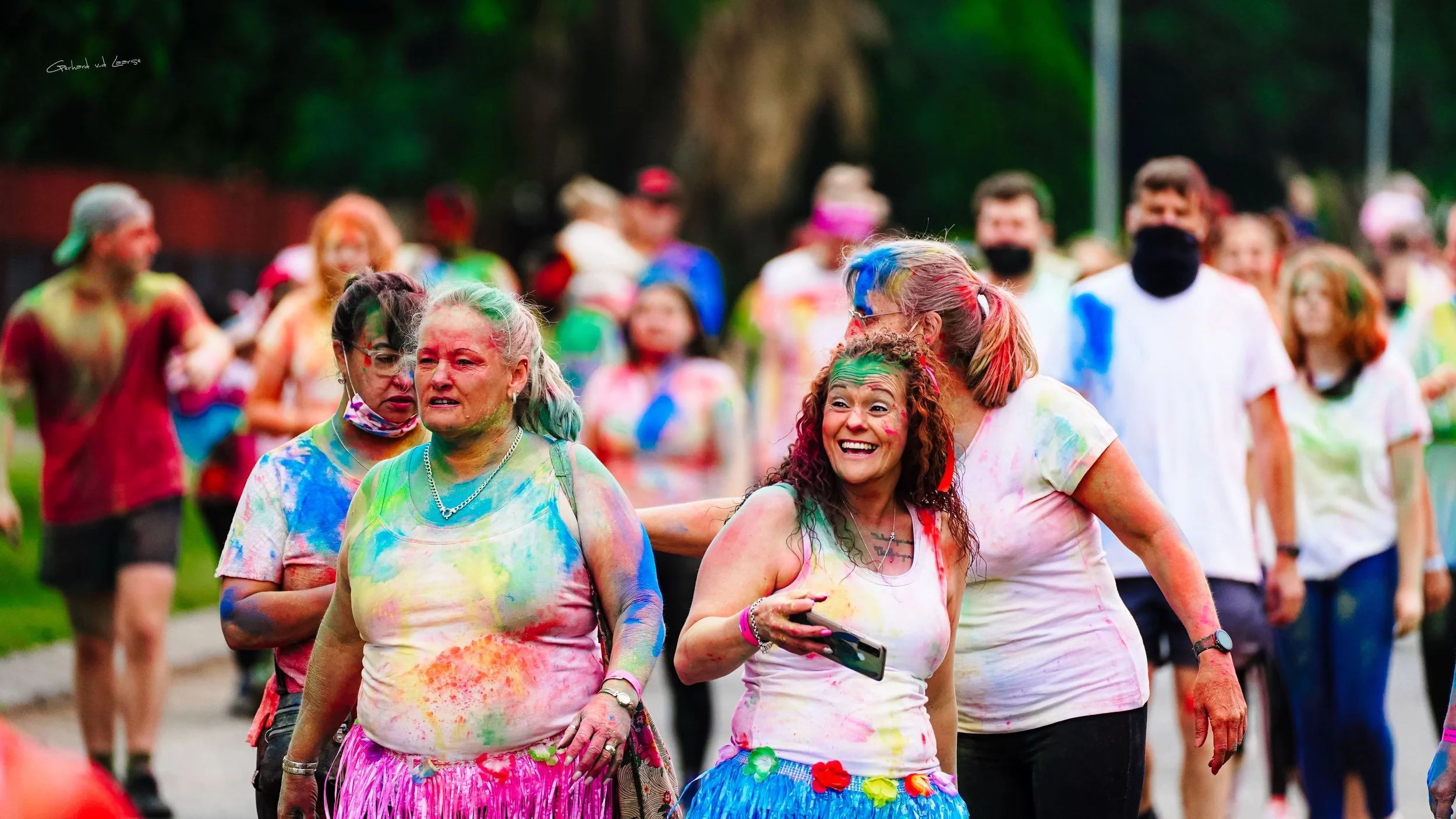 People celebrating at a color festival, covered in colorful powder, with smiling and joyful expressions, outdoors with trees in the background.