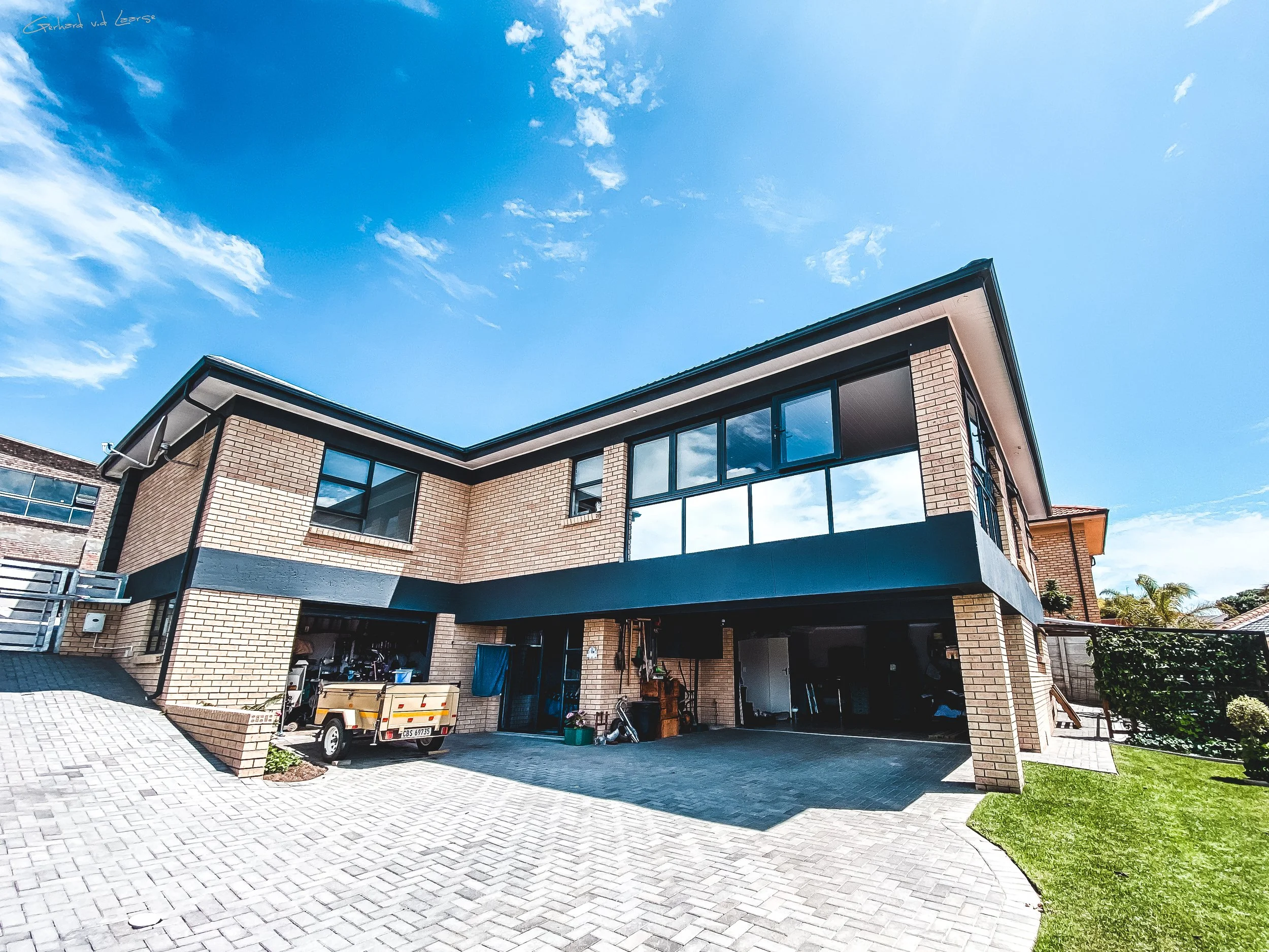 Modern two-story brick house with large glass windows, a paved driveway, and a small grassy area in front under a blue sky with clouds.