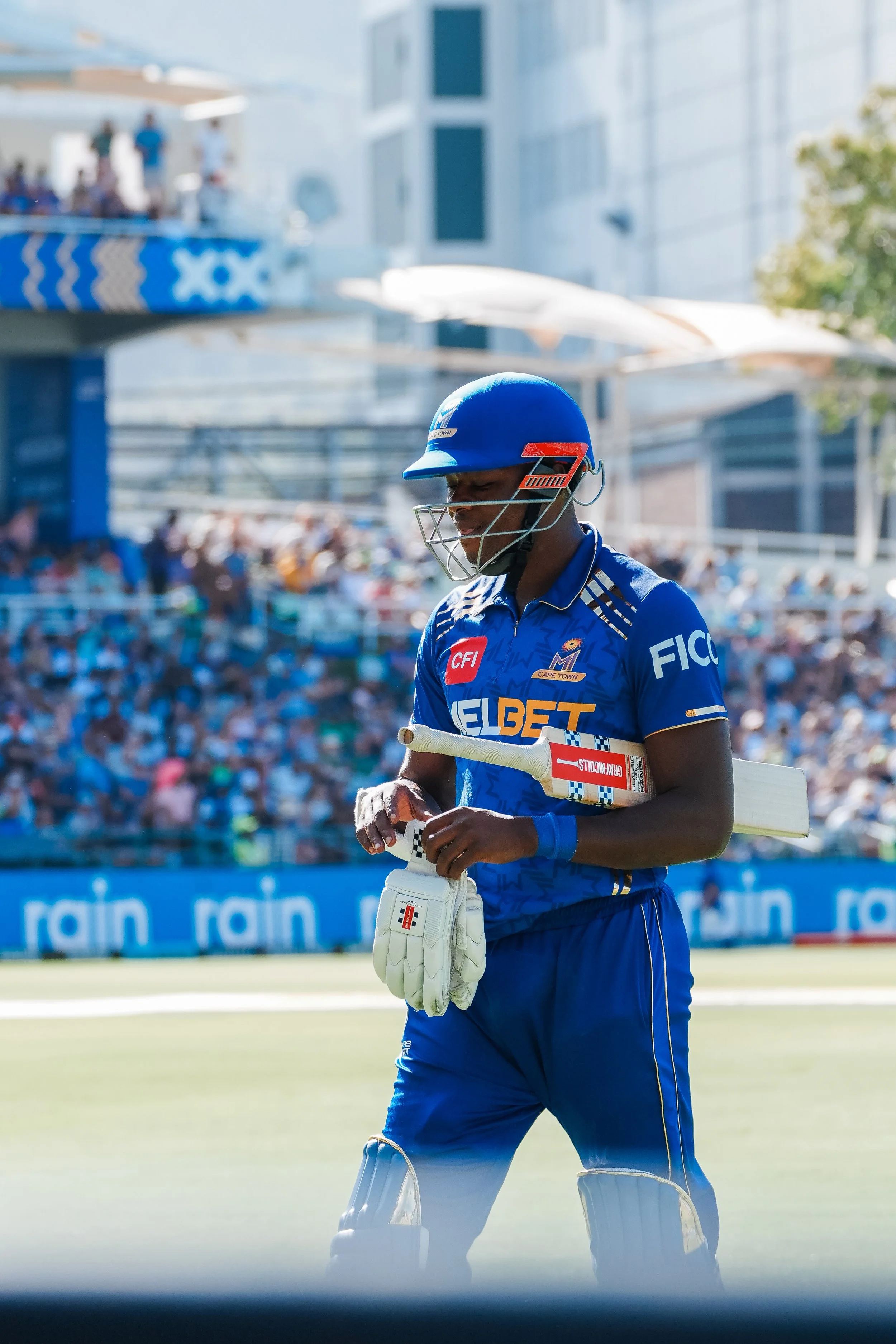 A cricket player in blue uniform and helmet holding a bat on the field during a match, with an audience in the background.