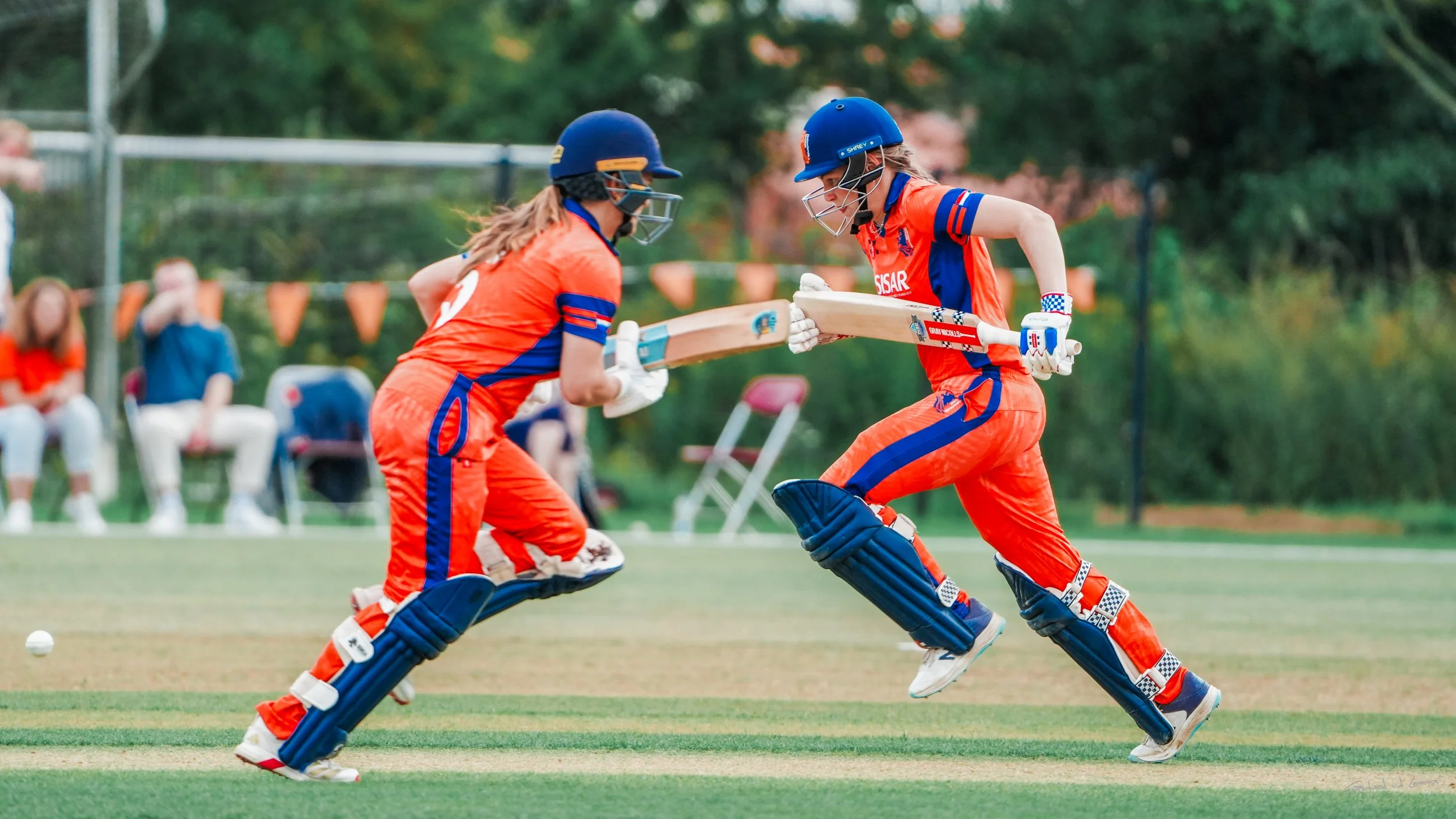 Two female cricket players in orange uniforms and blue helmets are engaged in a match, running towards each other on the field.