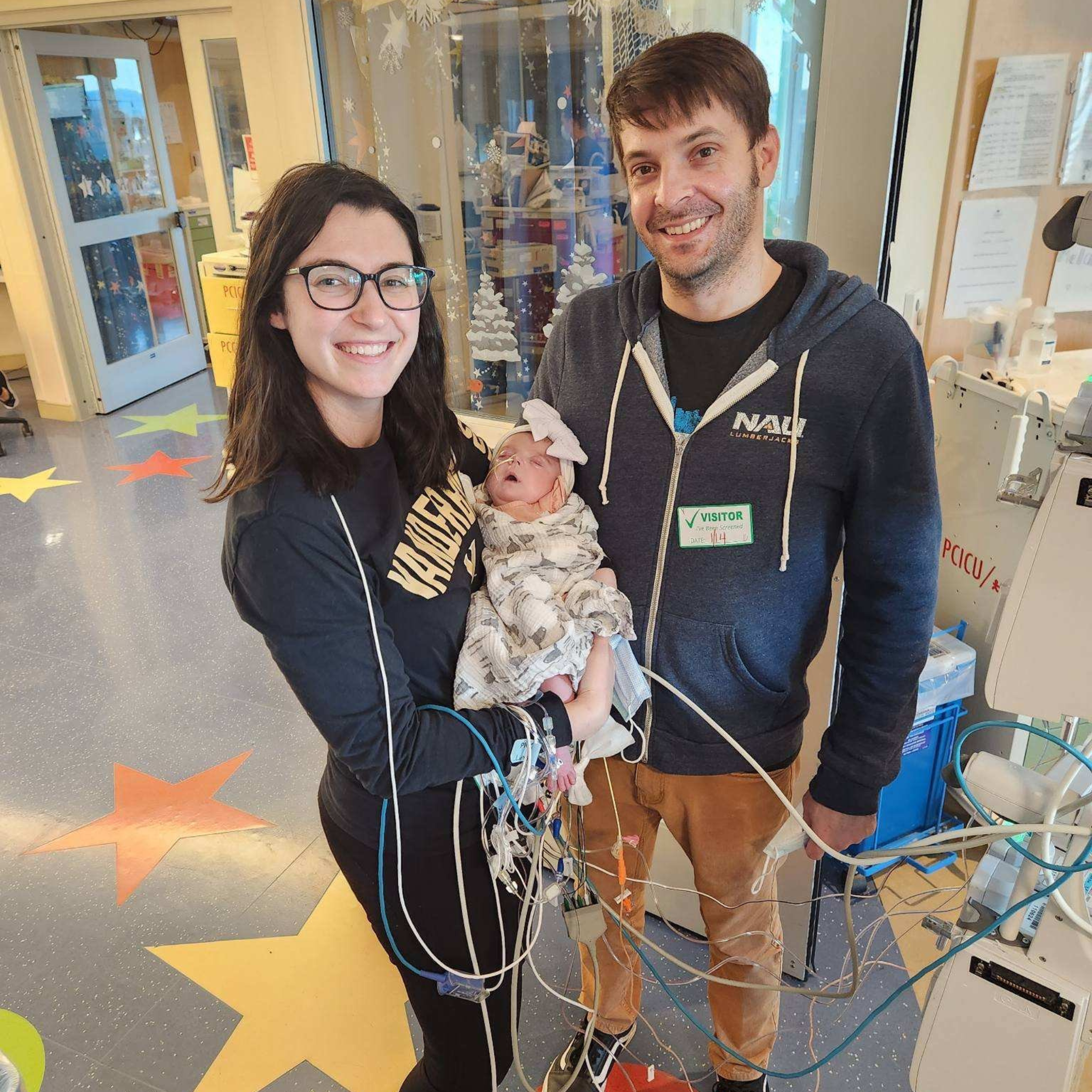 Young woman with glasses smiling at the camera while holding an infant, standing next to a man with a visitor badge in a hospital or medical facility. They are surrounded by medical wires and equipment with festive holiday decorations on the glass behind them.