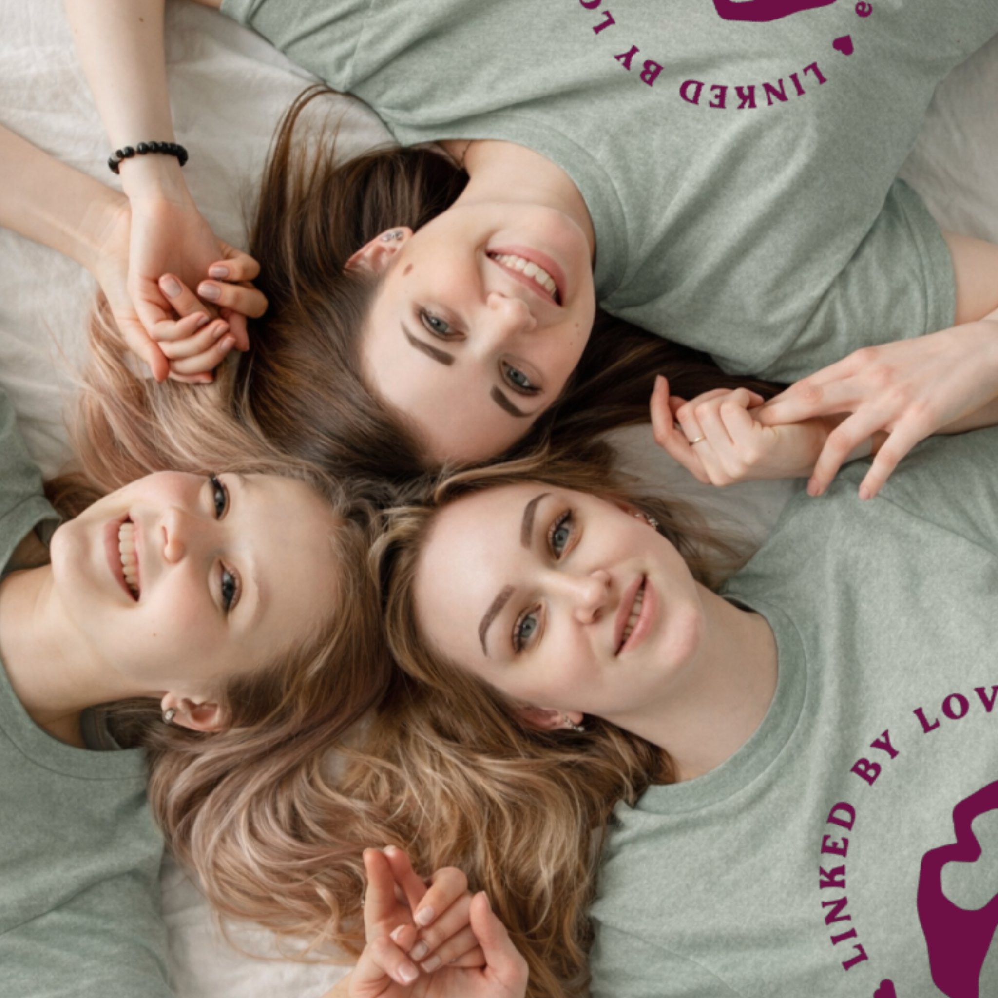 Close-up of two young women lying on the floor, smiling and making peace signs with their hands. One has long, wavy, light brown hair and the other has straight, dark brown hair. They are wearing casual clothing, with one in a grey shirt with purple text.