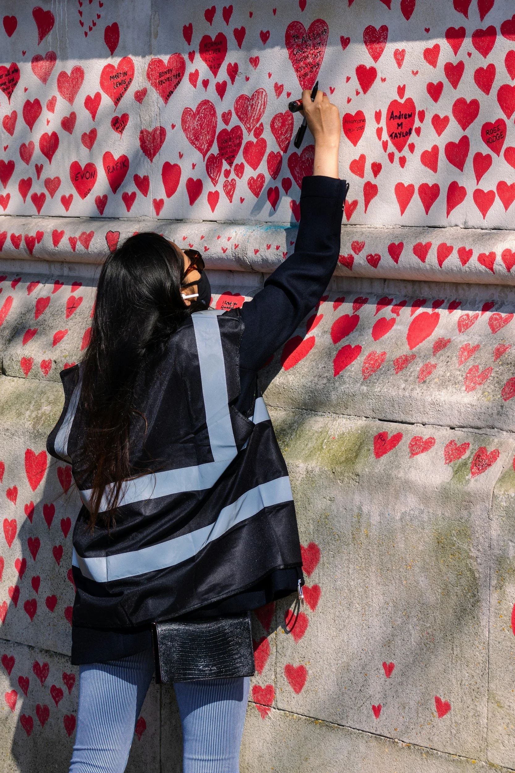 A woman standing in front of a wall covered in red hearts, writing on one of the hearts with a black marker. She is wearing a black and gray jacket, light blue striped jeans, and sunglasses. The wall has numerous hearts with some containing names and dates.