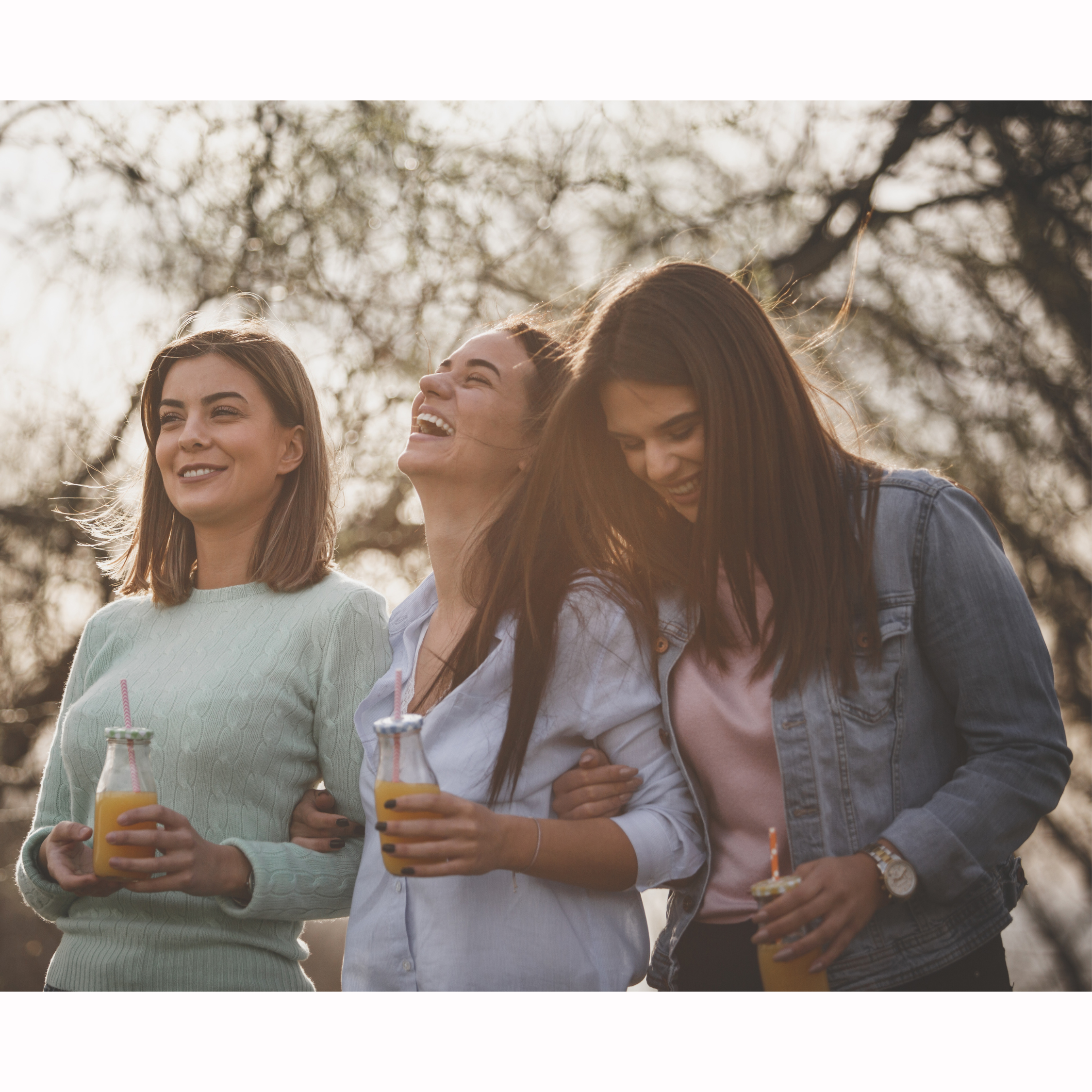 Close-up of three women outdoors holding orange drinks with straws, smiling and laughing with tree branches and sunlight in the background.