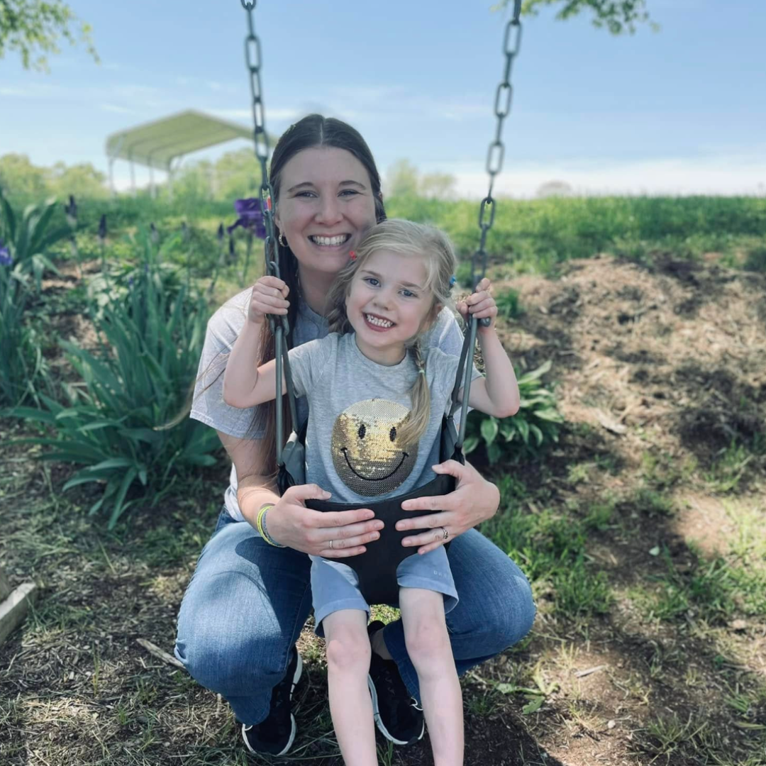 A woman and a young girl smiling and enjoying time on a swing outdoors in a garden or park.