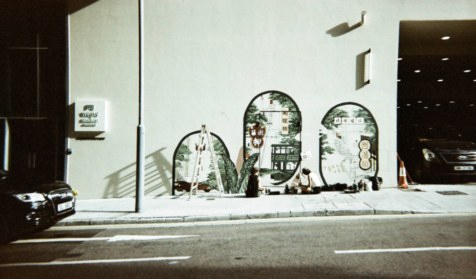 Two people sitting on the sidewalk in front of a mural painted on a white wall, showing illustrations of plants and traditional Chinese symbols, with a ladder beside them and a small traffic cone nearby.