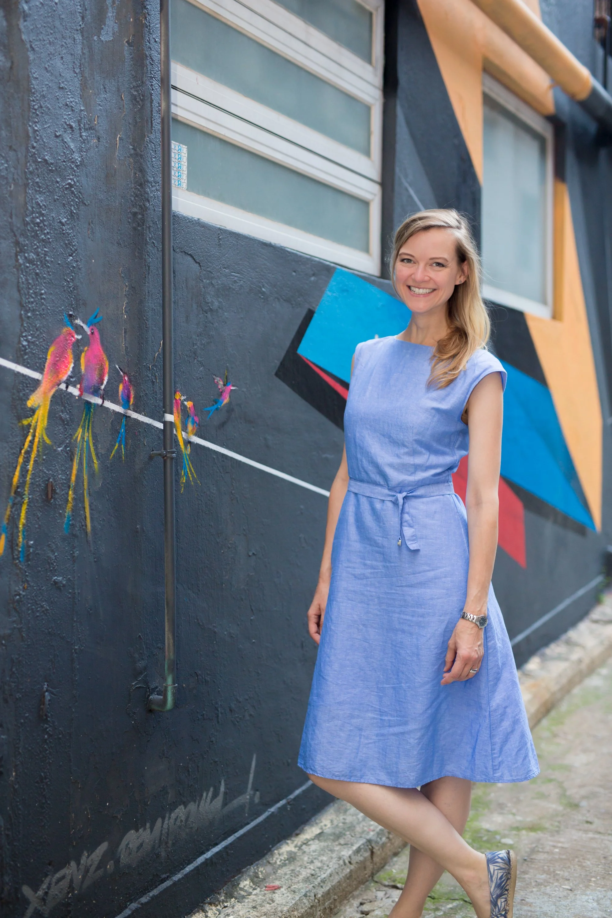 A woman in a blue dress standing next to colorful wall art of birds and geometric shapes.