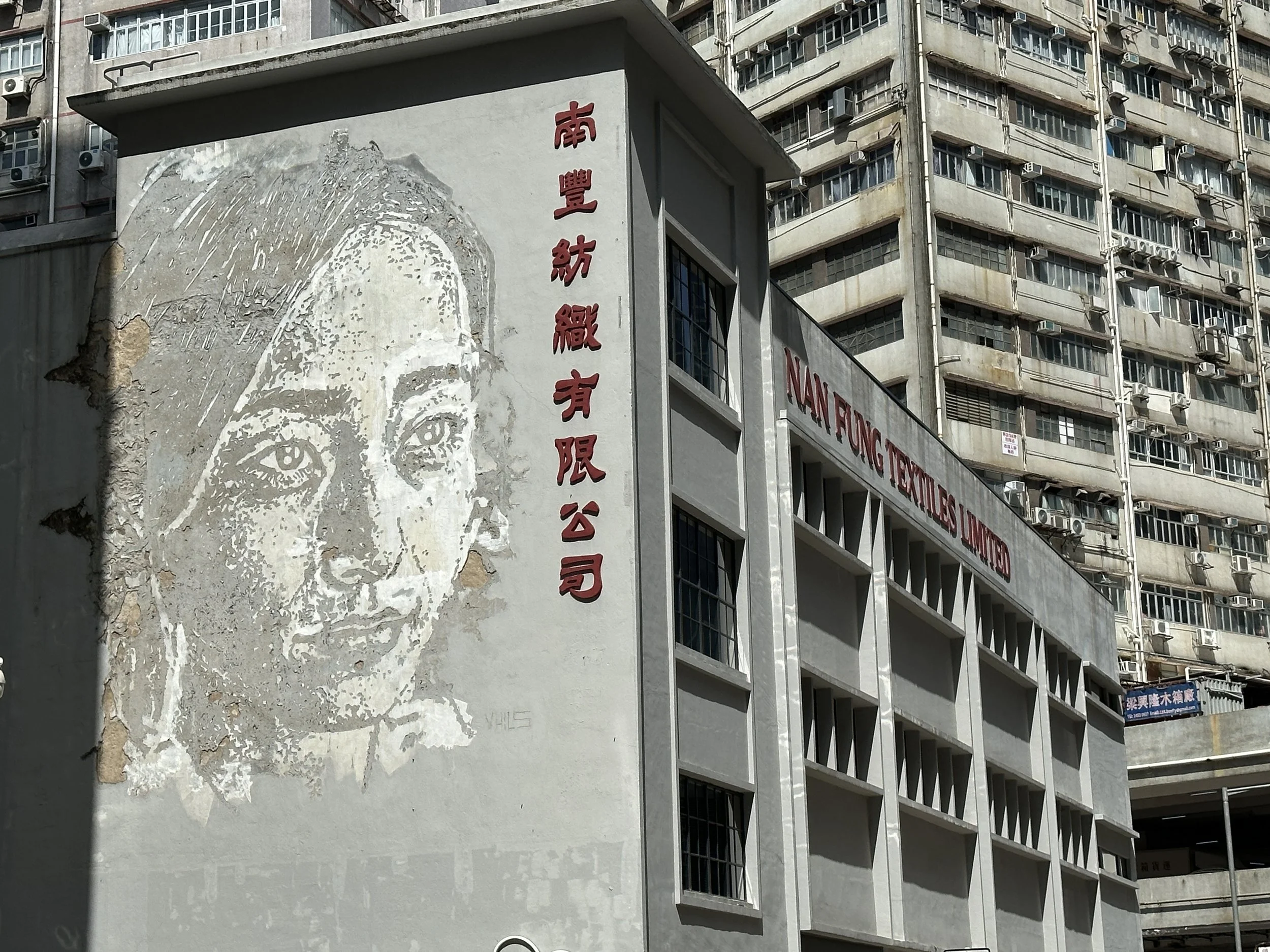Street view of a building with a large mural of a woman's face and Chinese characters on the side. The building has white walls, multiple windows, and is labeled 'NAN FUNG TEXTILES LIMITED'.
