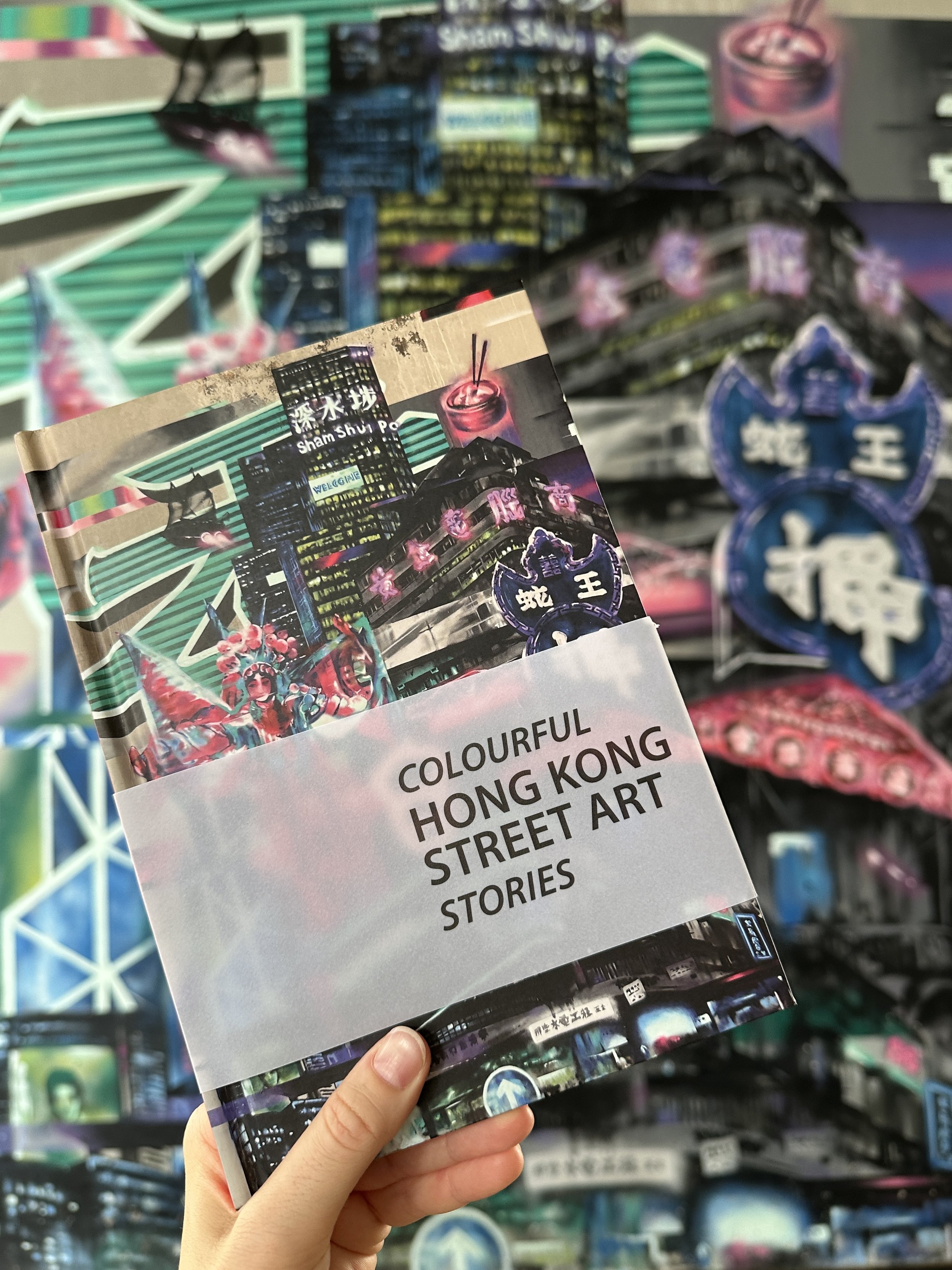 The book 'Colourful Hong Kong' held against the iconic street art mural by Sinic Choy in Sham Shui Po, Hong Kong.