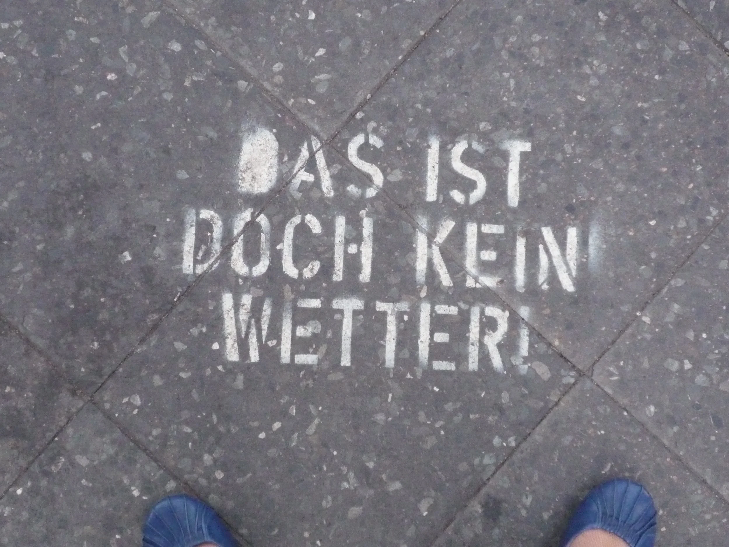 The quote 'Das ist doch kein Wetter' in bold white spray paint on a grey Berlin sidewalk. Captured in 2010 by Alex, author of Colourful Hong Kong – Street Art Stories and founder of Wanderlust Walks.