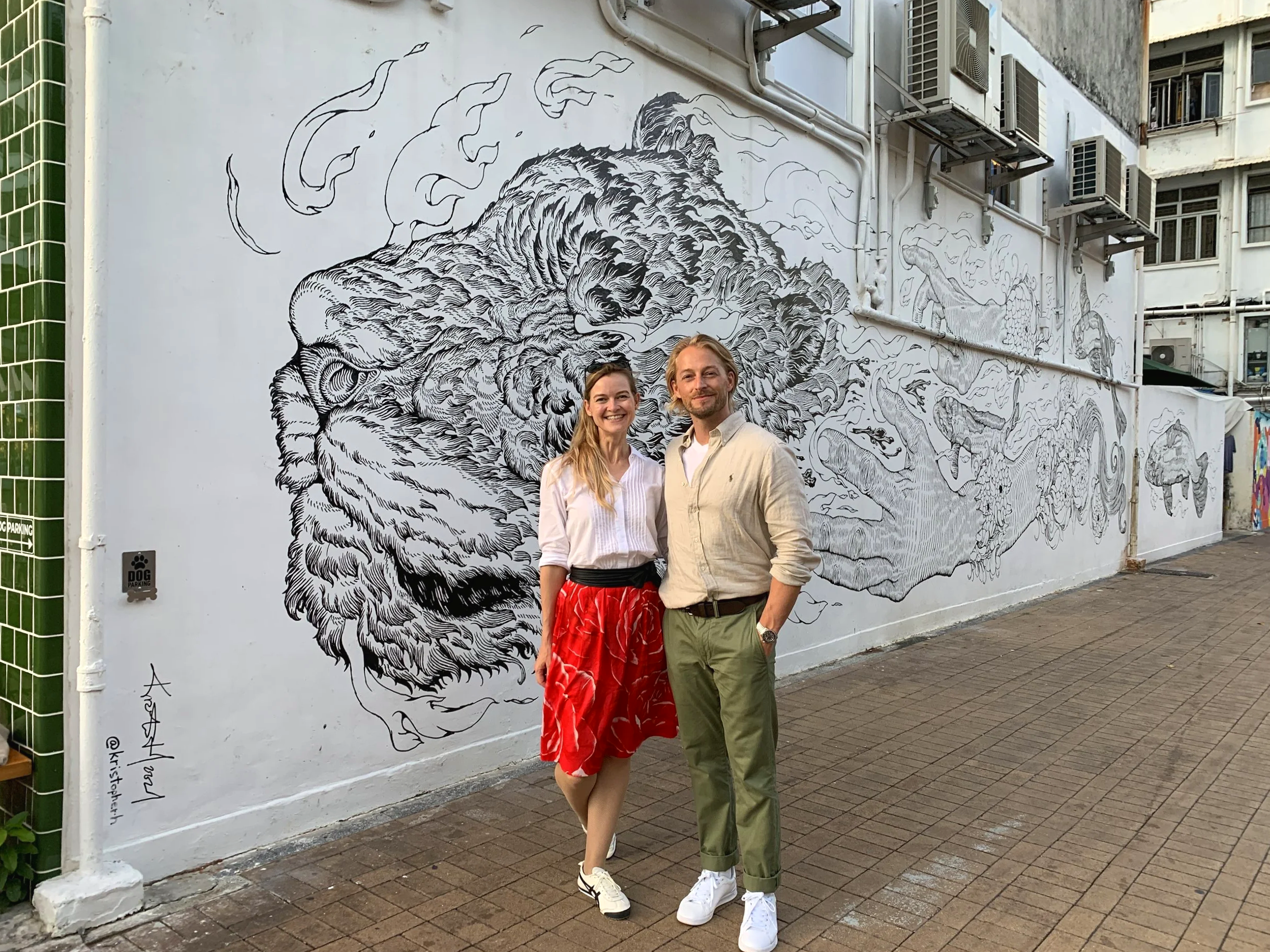 A man and woman standing in front of a large mural of a tiger on a white wall. The mural is detailed and black and white, with the tiger's face appearing in a stylized, artistic manner.