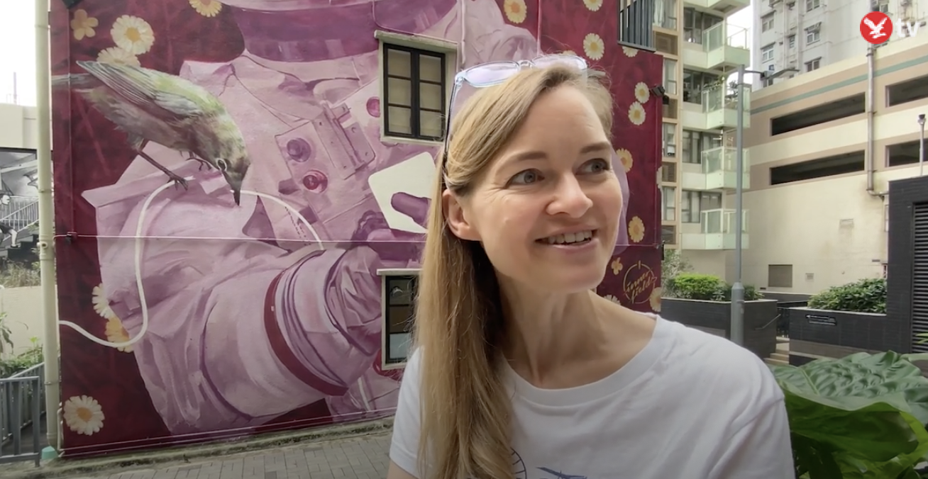 A woman with long blonde hair smiling, standing outdoors near a colorful mural featuring a bird, daisies, and abstract shapes, in an urban setting.