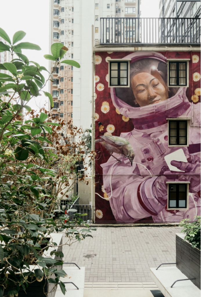 Mural of a woman in a spacesuit holding a flower and a smartphone, with a bird perched on her arm, on an urban building wall surrounded by plants.