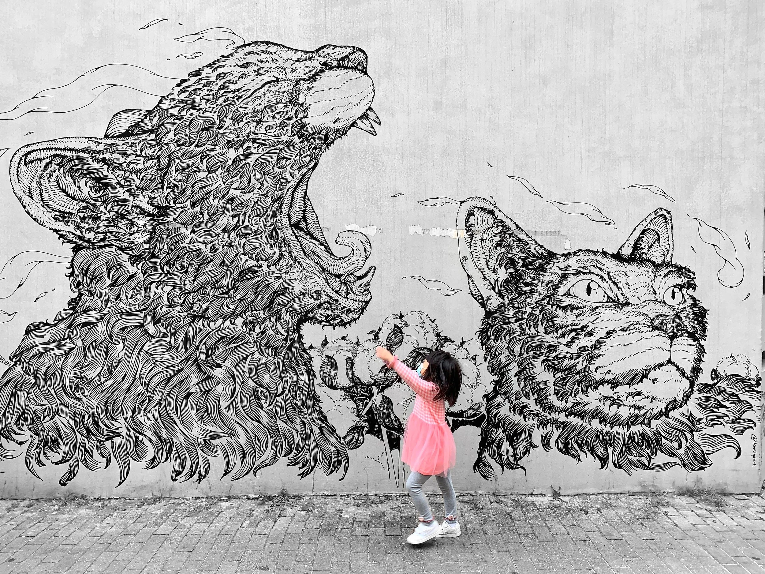 A young girl in a pink dress and gray leggings is standing in front of a wall mural of a roaring lion and a calm cat, both depicted with detailed black lines on a gray background.
