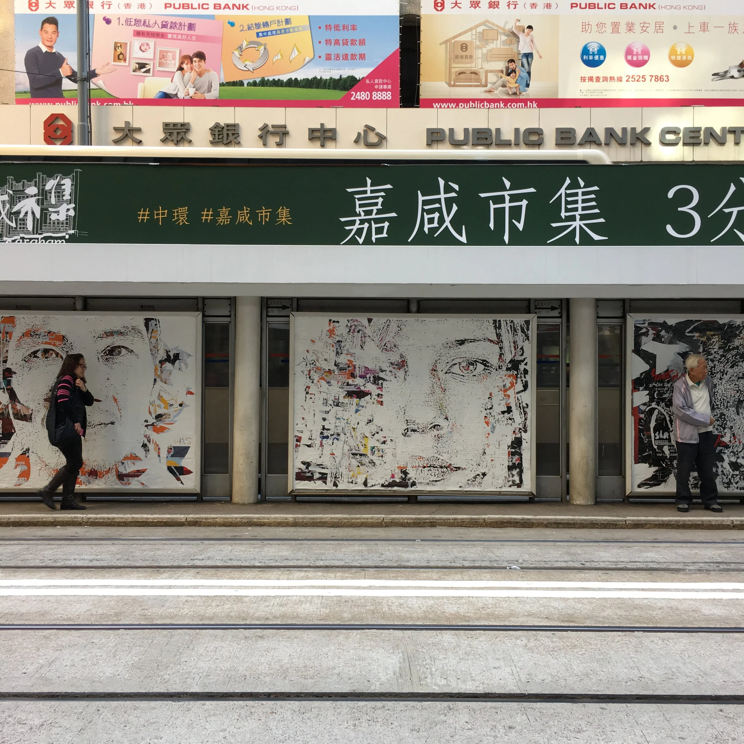 Street scene in Hong Kong featuring large posters with digital art of faces, a public bank sign, and a green sign with Chinese characters, with pedestrians walking along the sidewalk.