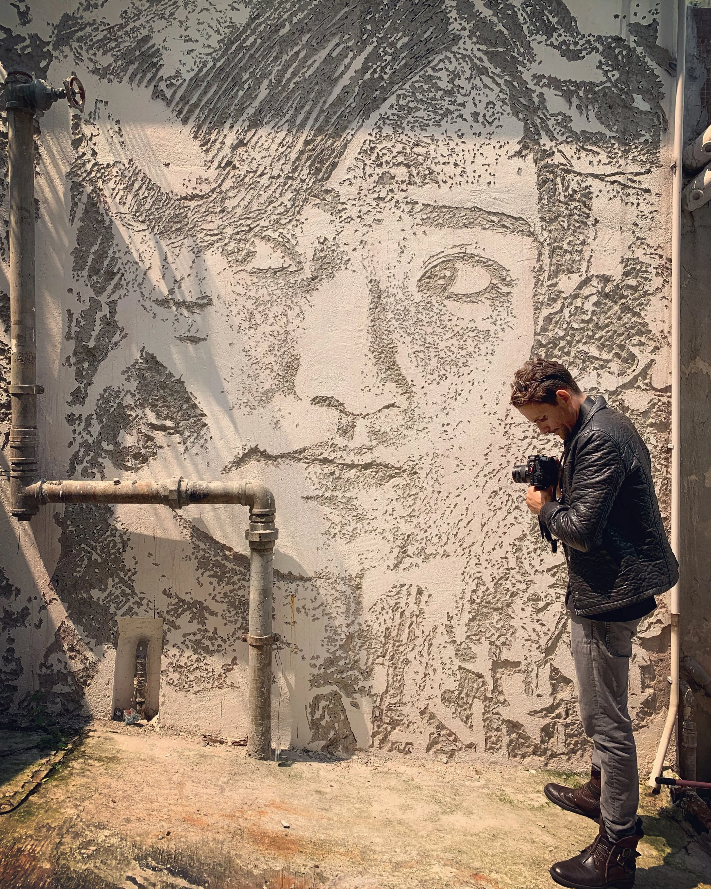A man in a black leather jacket and dark pants is standing outside, looking down at a camera he is holding in his hands. Behind him is a large mural on the wall, depicting a detailed, stylized portrait of a person's face with closed eyes, created using textured, carved patterns. There are also some pipes on the wall near the mural.