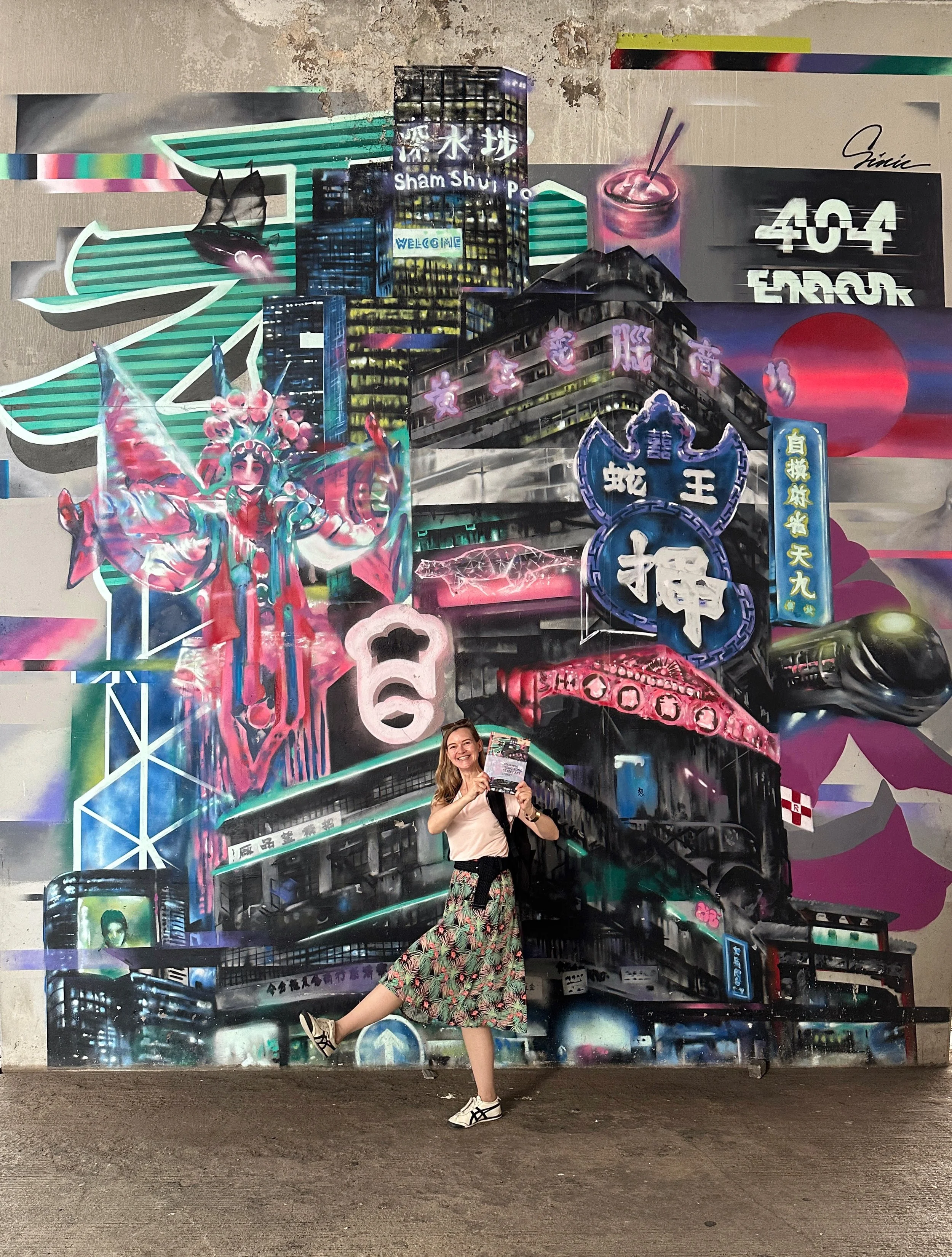 A woman smiling and holding a brochure or flyer while standing in front of a colorful urban graffiti mural featuring cityscape images, neon signs, Asian characters, and a traditional opera or theater performer, creating a vibrant, artistic scene.