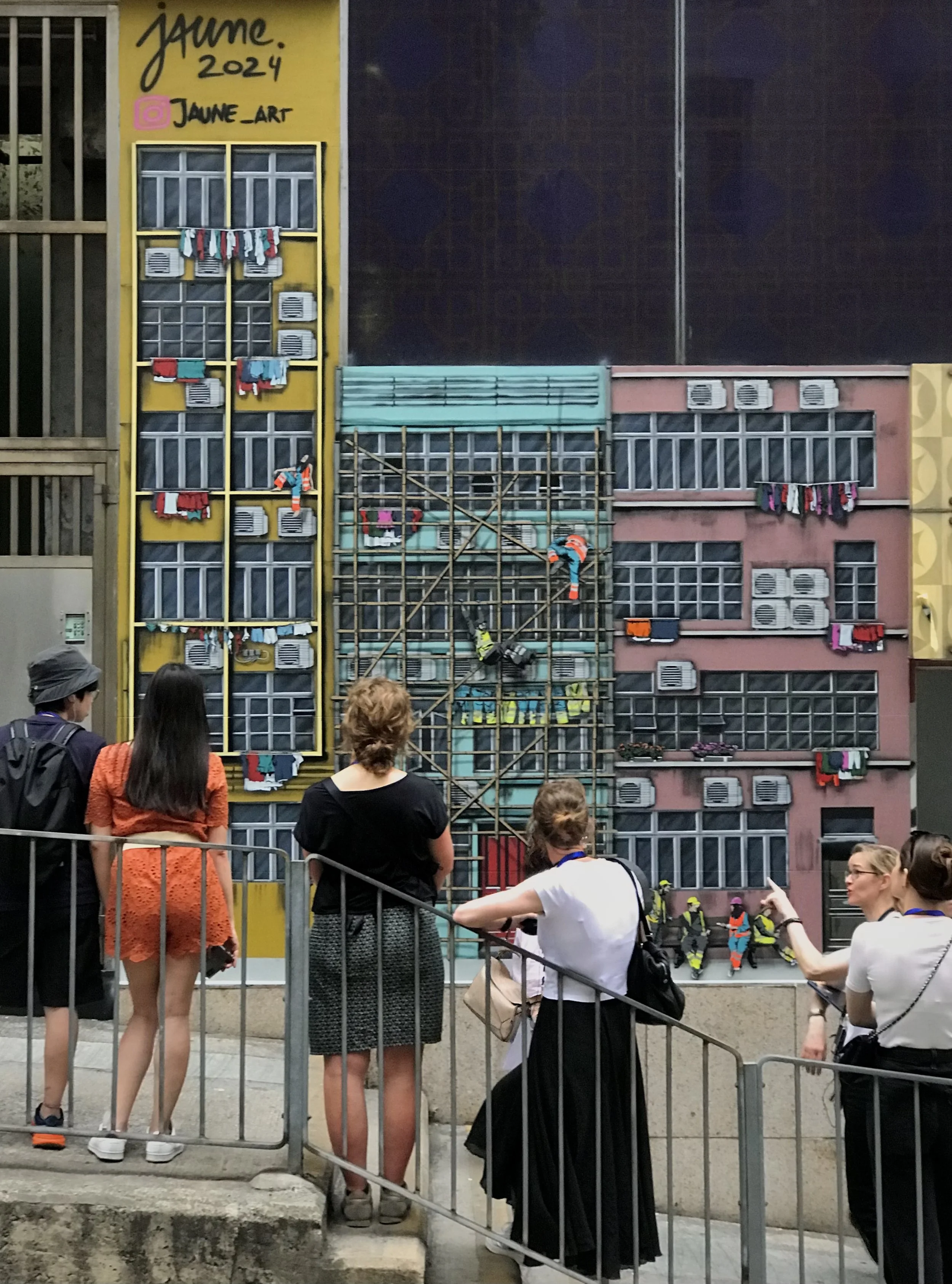 A group of people watching a staged construction scene on a colorful building facade. The facade features painted apartment windows with hanging laundry and wall-mounted air conditioning units. Several actors dressed as construction workers are on scaffolding and on the ground, acting out a construction scene.