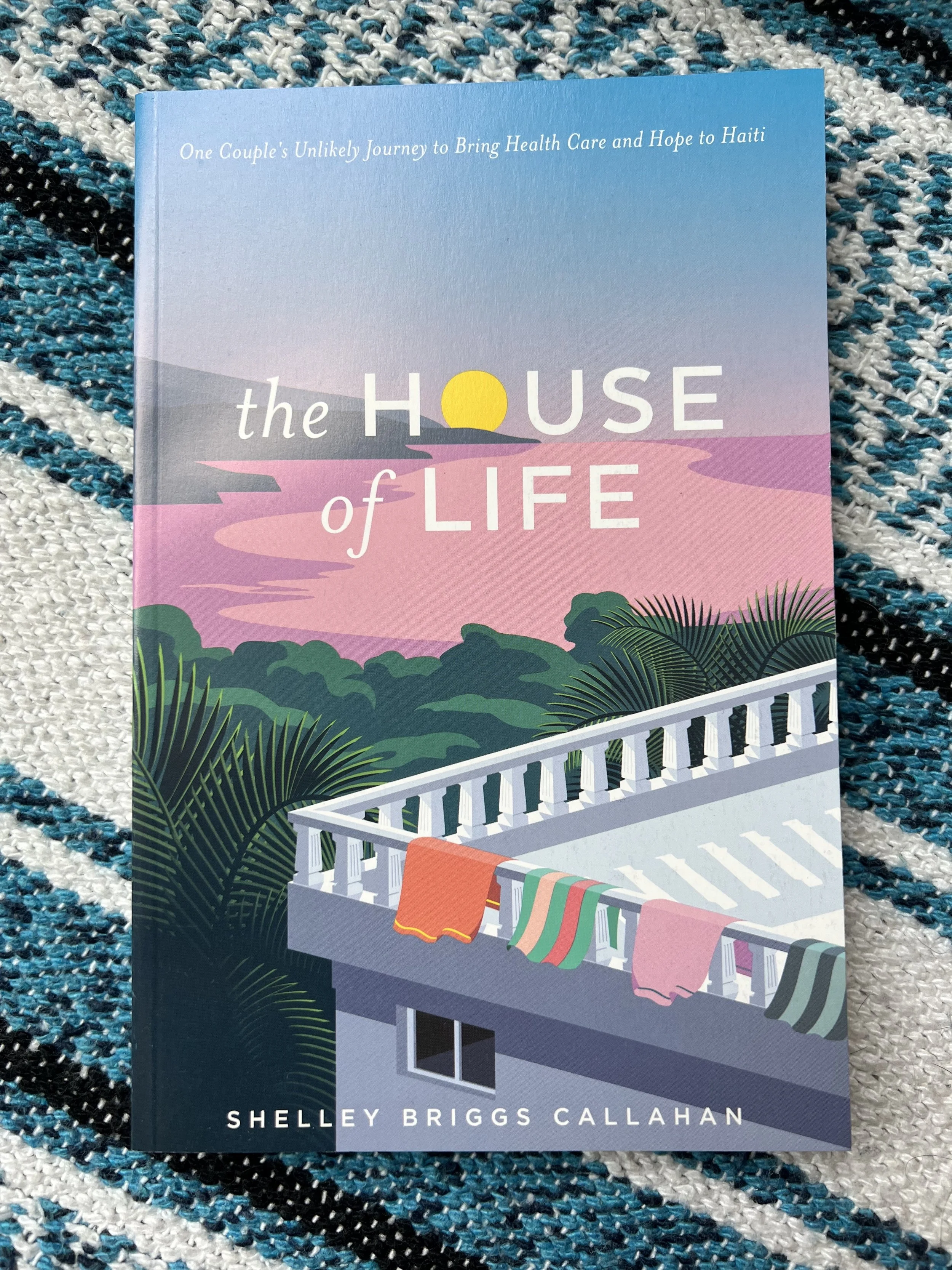 A book titled 'The House of Life' by Shelley Briggs Callahan, featuring an illustration of a balcony with colorful towels hanging and surrounded by palm trees, with a sunset in the background.