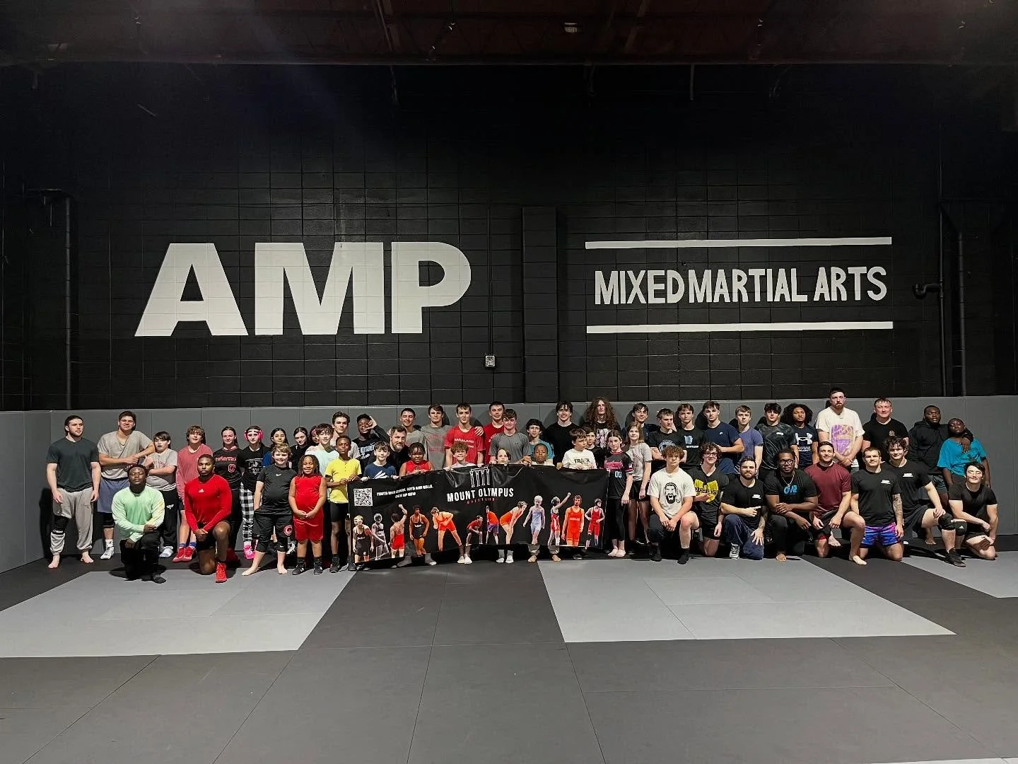 Group of children and adults in a martial arts gym, some holding a banner that reads, 'Mount Olympus.' The gym has black walls with large white lettering that says 'AMP' and 'Mixed Martial Arts'.