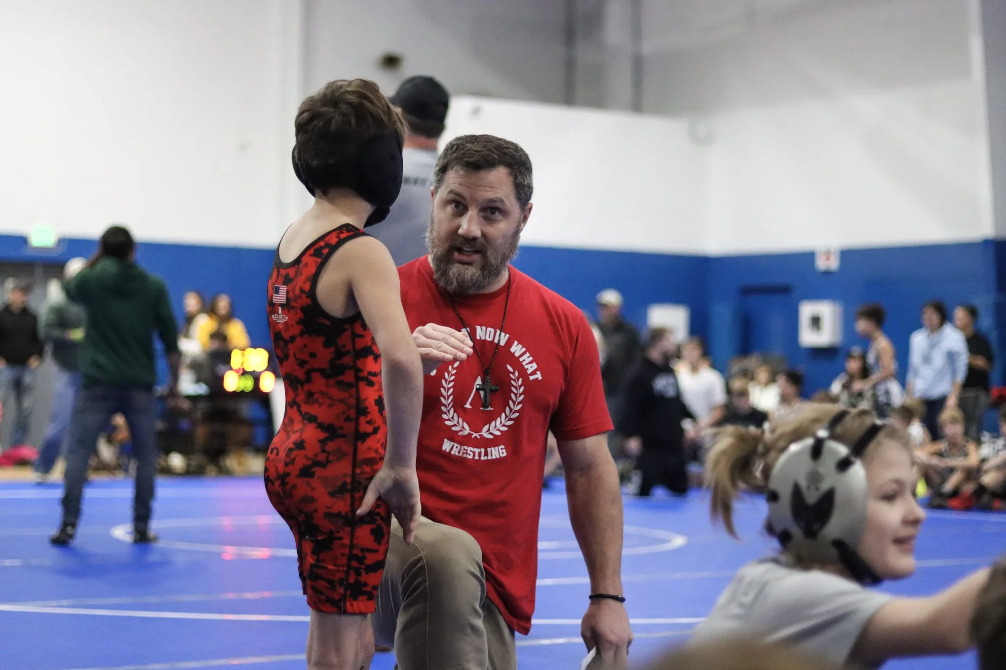 A man talking to a young girl in a wrestling gym, with several other people in the background, some sitting and some standing, and a blue wrestling mat on the floor.