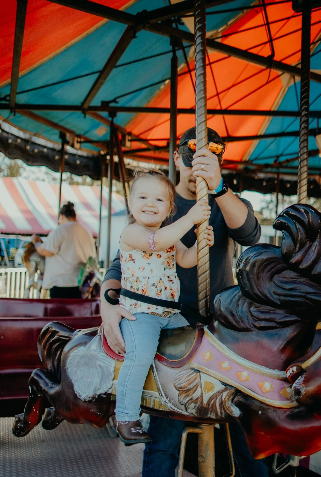 A young girl riding a carousel horse, smiling at the camera, holding onto the pole while a man, wearing sunglasses and a cap, is holding her securely. The carousel is under a colorful blue and orange canopy, with other fair attendees and a striped tent in the background.