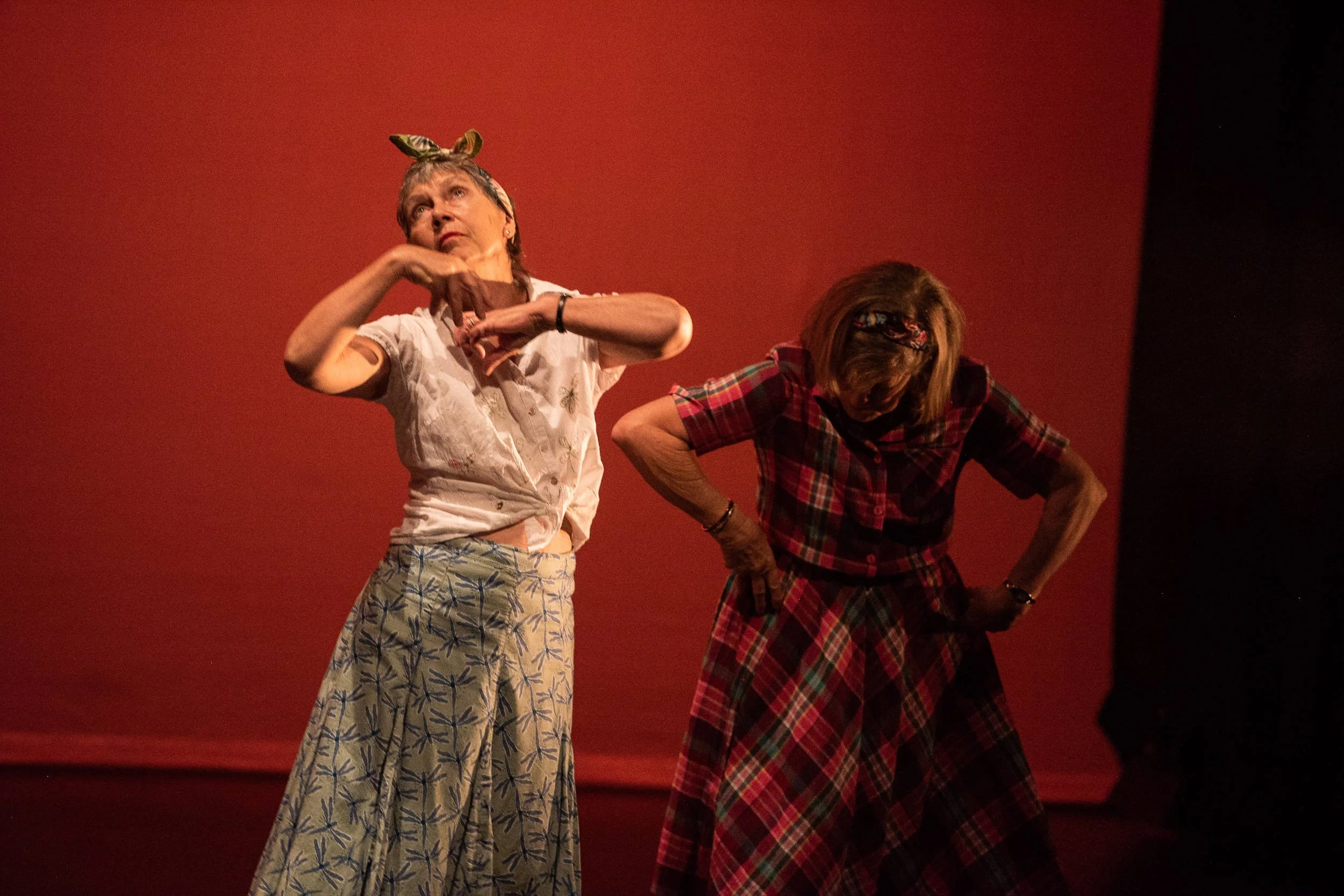 Two older women perform on stage against a deep red backdrop, one gazing upward with raised hands in a floral skirt and white blouse.
