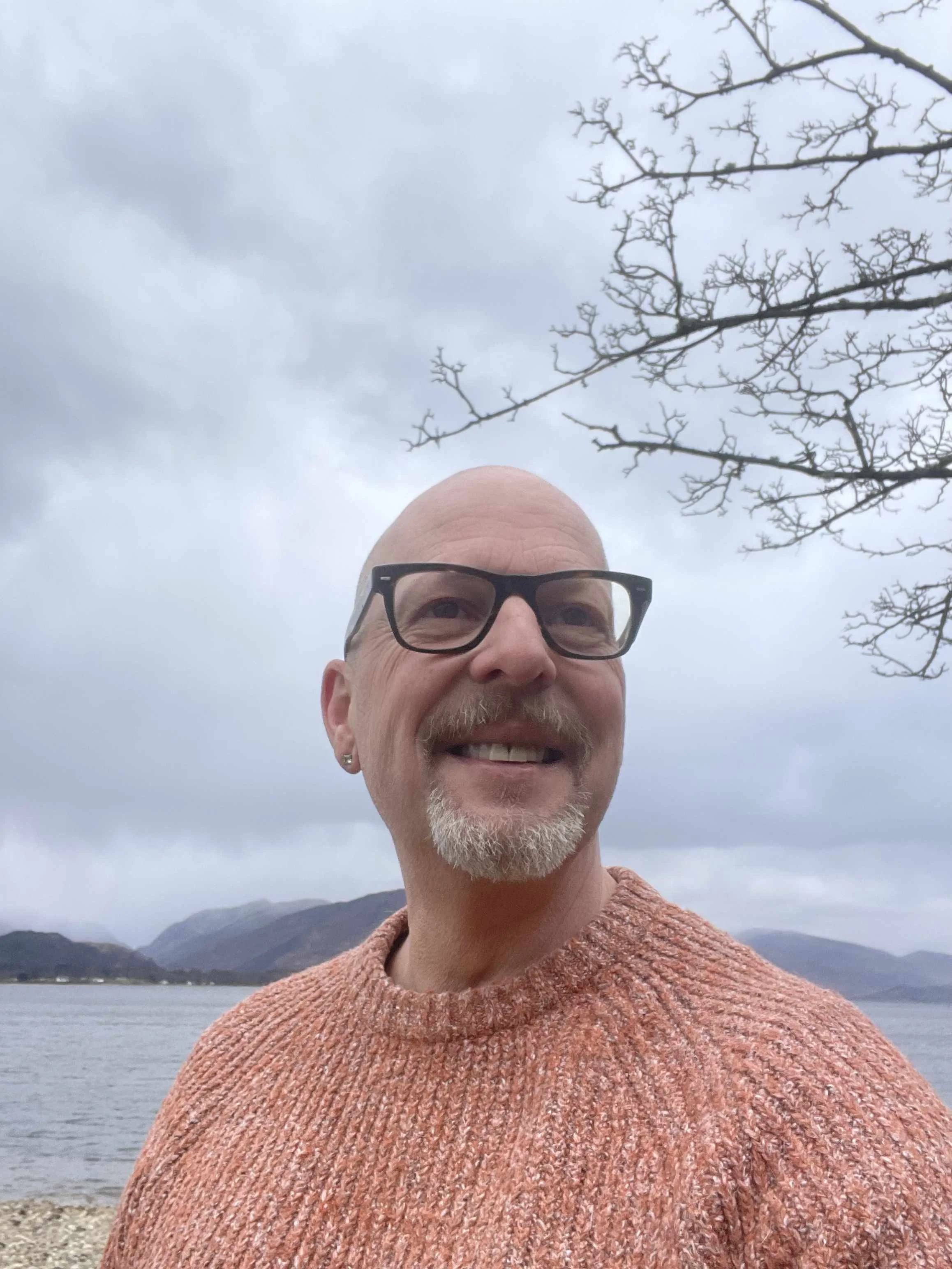 ohn Darvell, Artistic Director of NOCTURN Creative, smiling outdoors beside a Scottish loch with hills and overcast sky behind him.