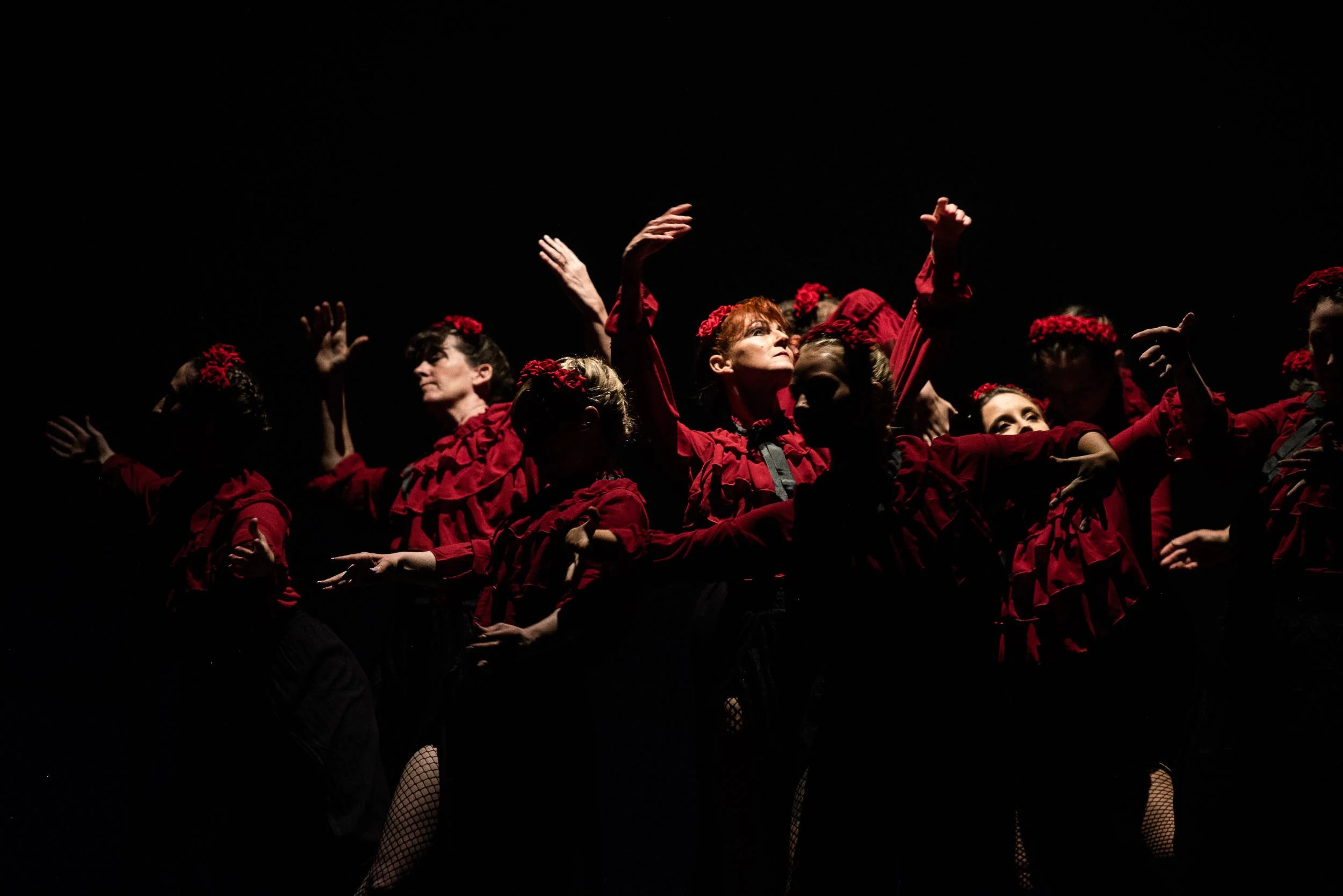 Large ensemble of performers in deep red ruffled costumes and flower headpieces raise their arms on a dramatically lit dark stage.