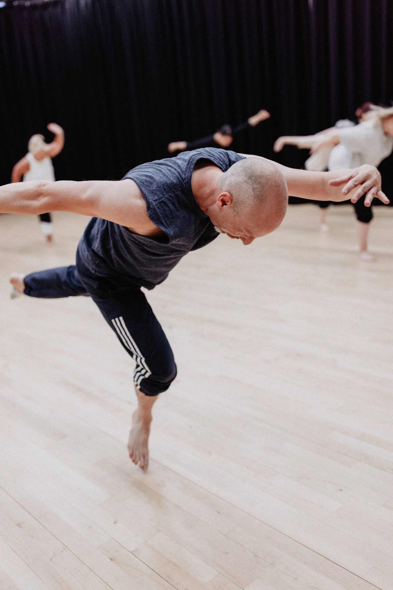 Bald male dancer balances on one leg with both arms extended wide in a dance studio, other participants moving in the background.