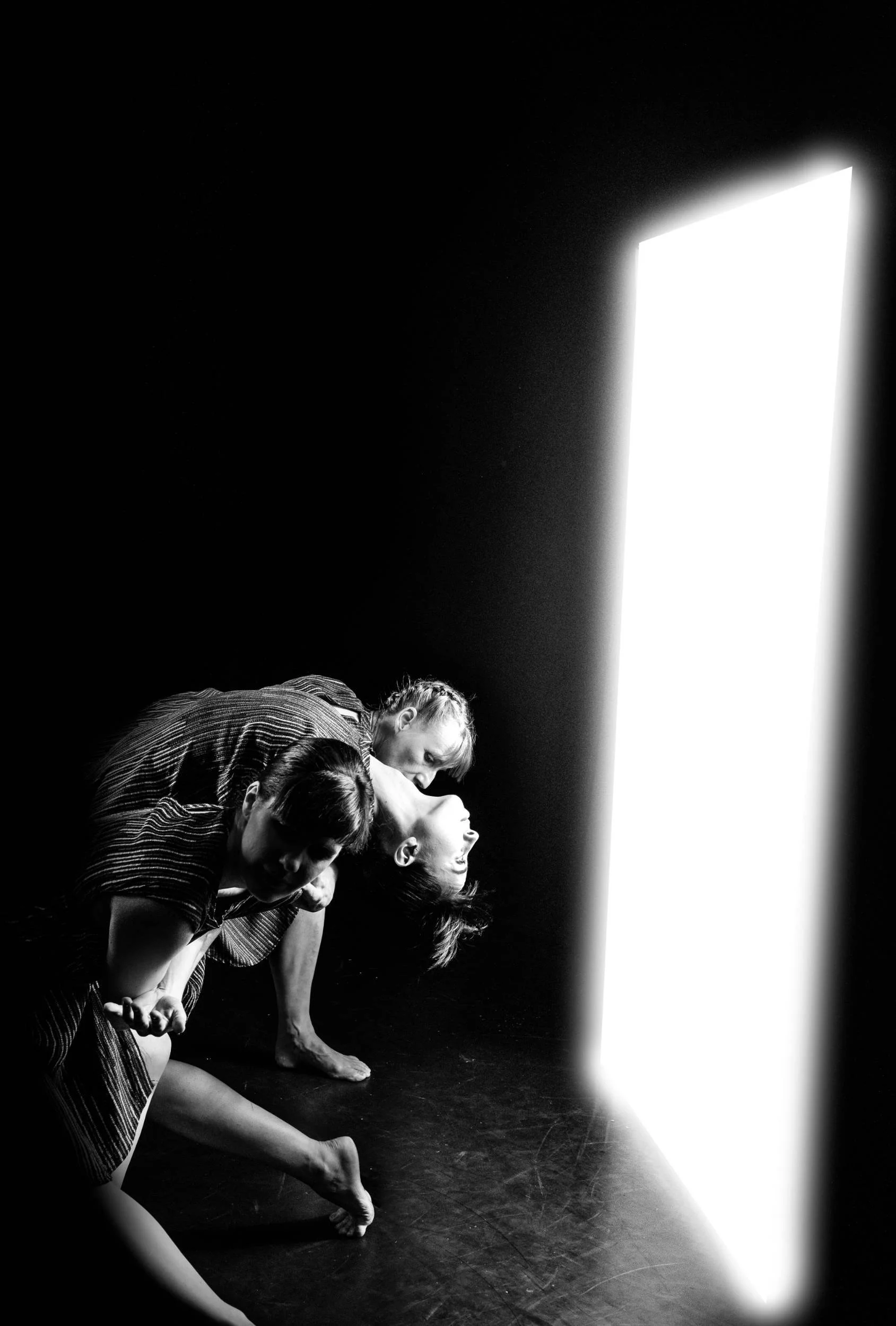 Three dancers in striped clothing bend low beside a tall vertical column of white light against a black stage, photographed in black and white