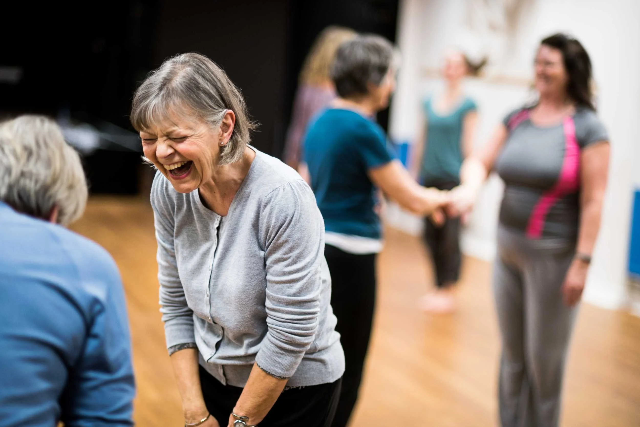 Older woman with grey hair laughs joyfully mid-movement during a dance workshop, with other participants visible in the background.