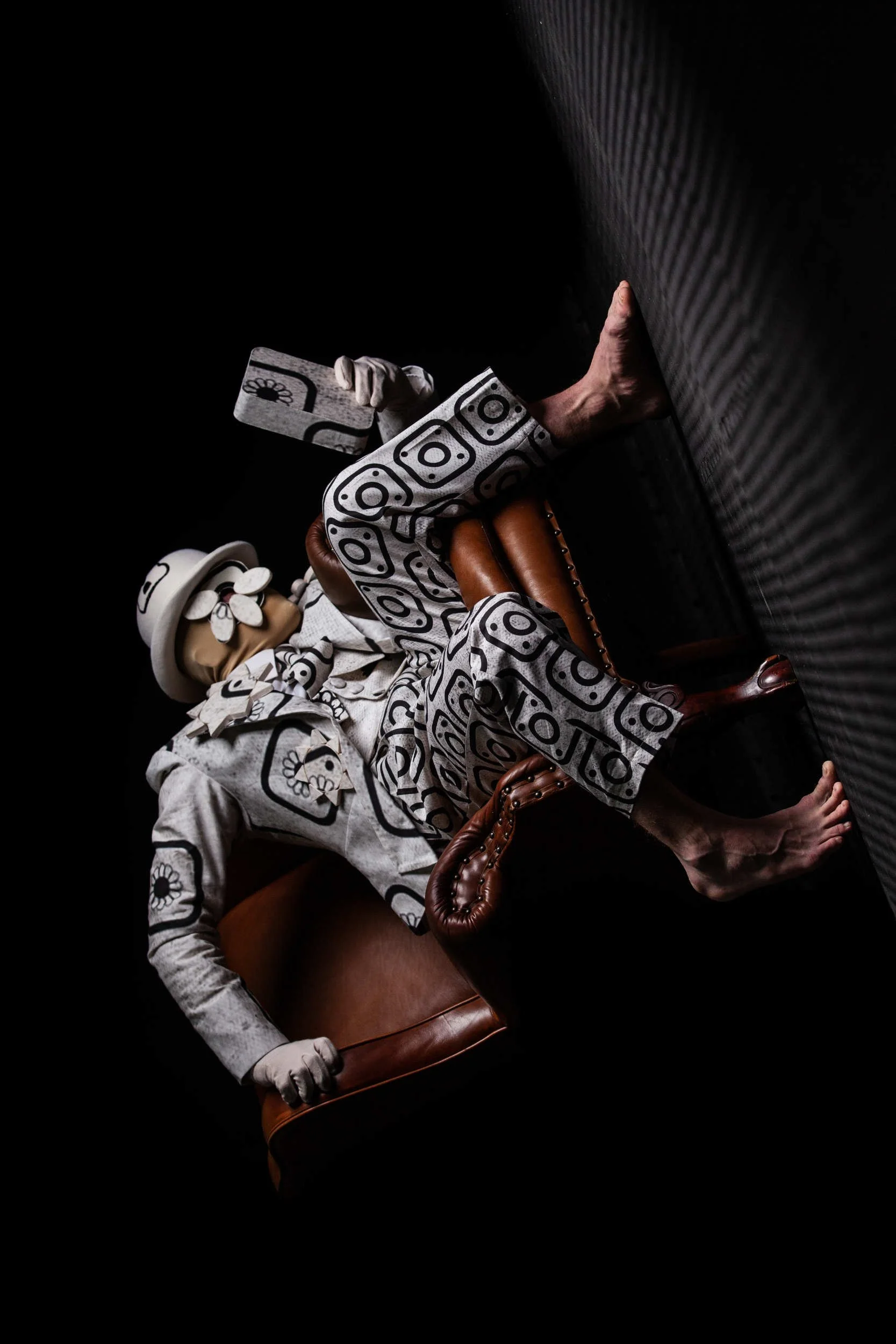 Surreal performance artwork by John Darvell for NOCTURN, featuring a costumed figure in a patterned suit reclining in a chair with legs pressed against a wall in dramatic low-key lighting.