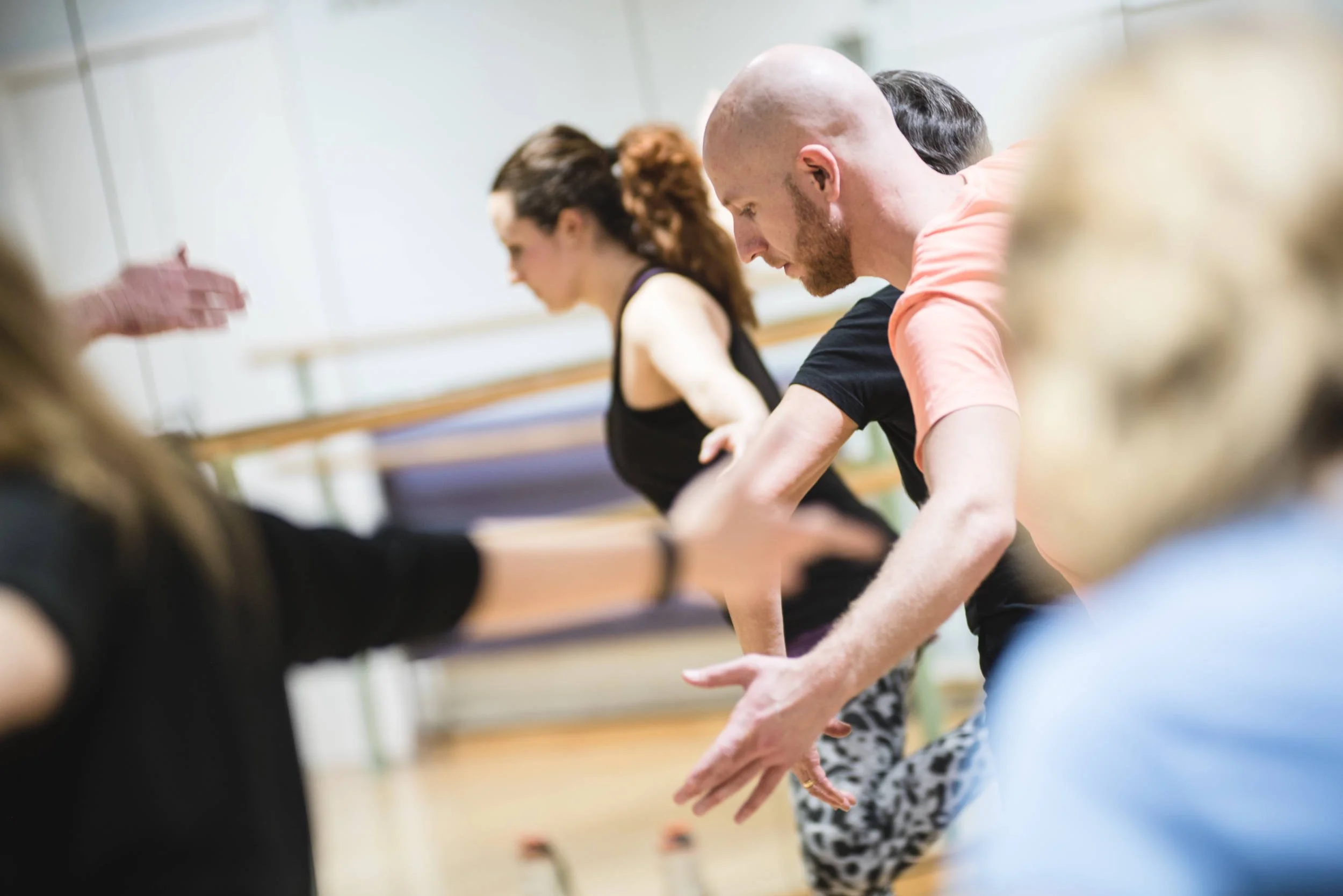Dancer in salmon top leans forward with arms extended during contemporary dance workshop in mirrored studio with ballet barre.